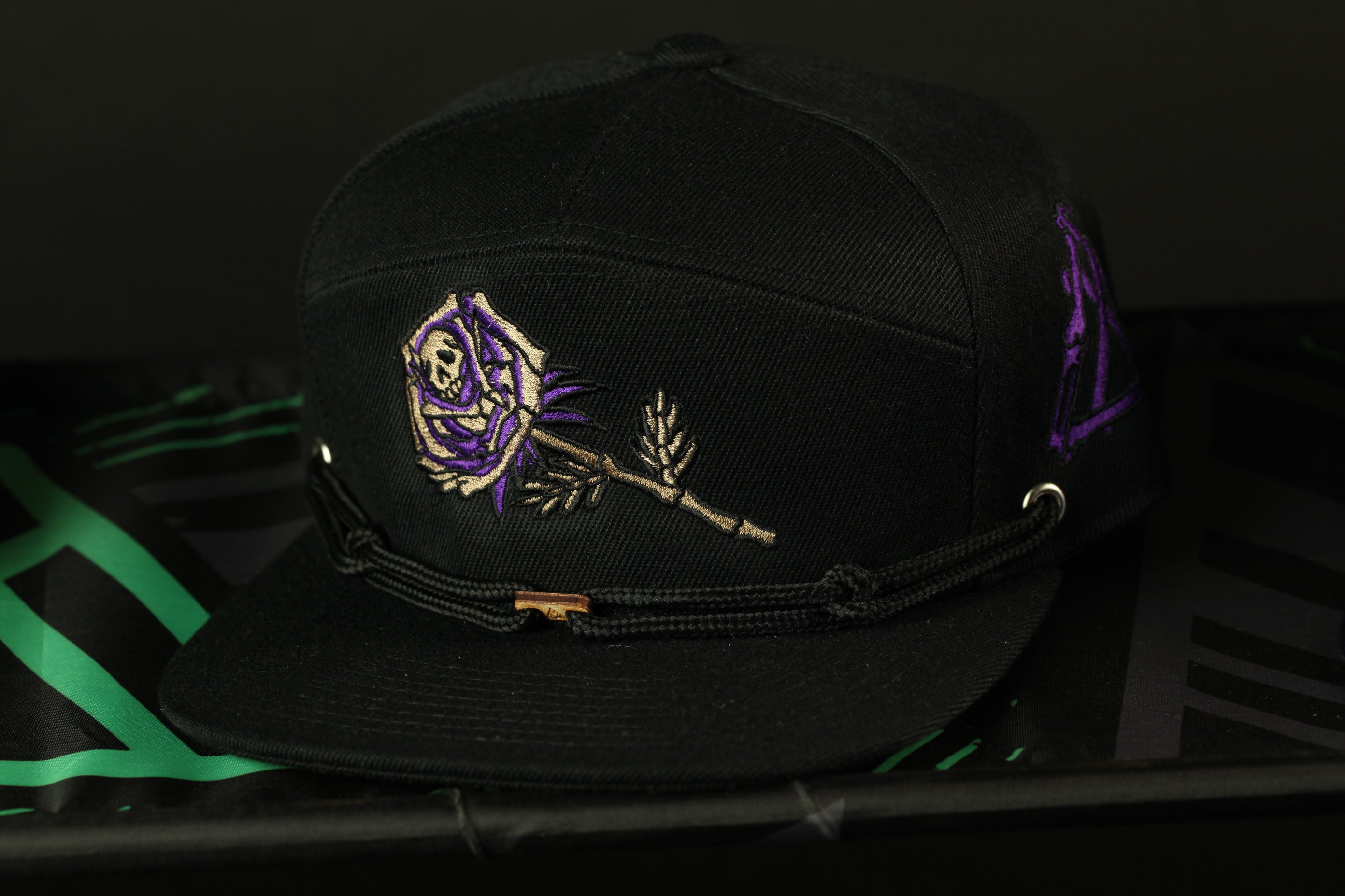 A black baseball cap with a purple rose embroidered on it, sitting on a black surface.