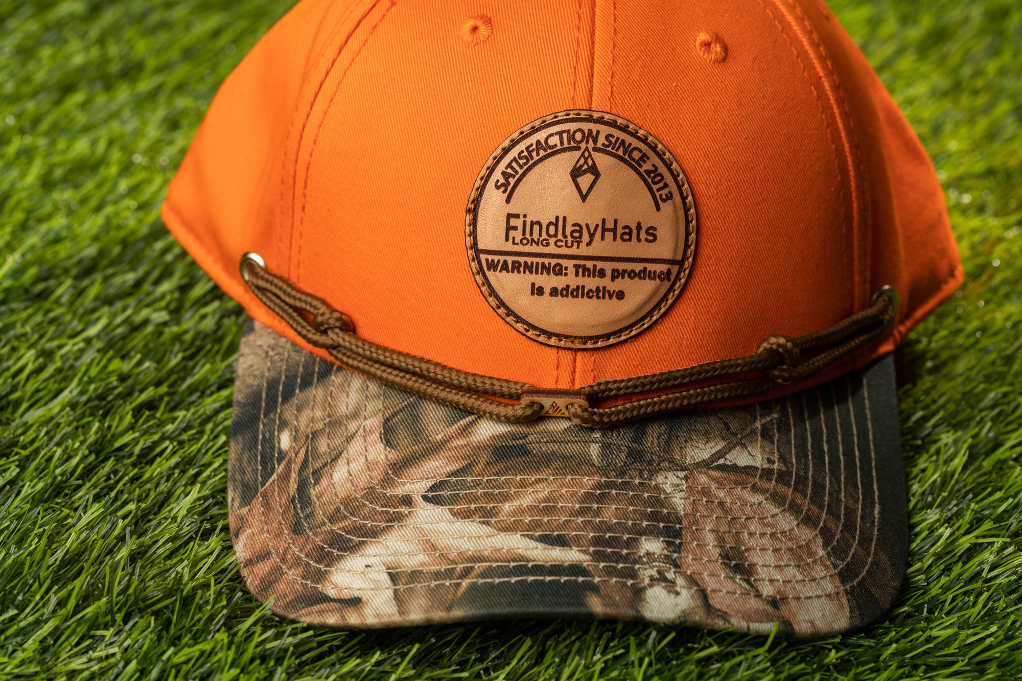An orange baseball cap with a camouflage pattern and a circular logo on the front, placed on a grassy surface.