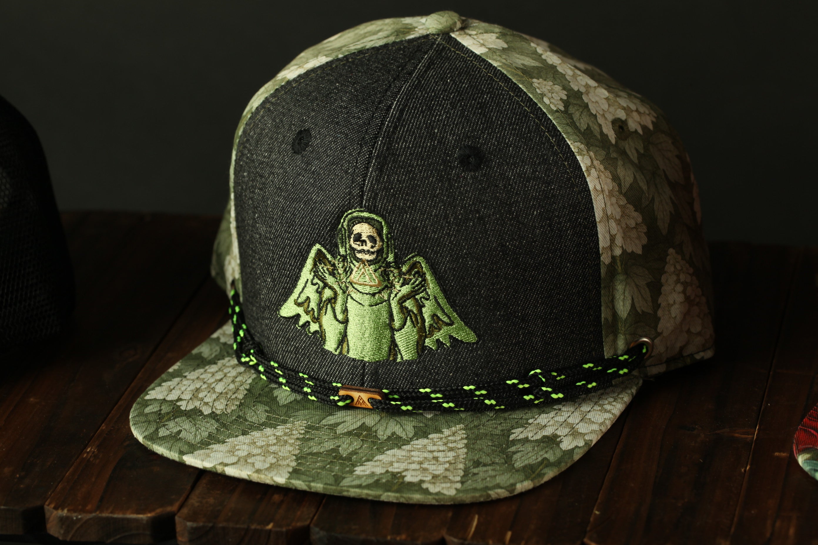 A camouflage baseball cap with a skull and angel embroidered on it, sitting on a wooden surface.