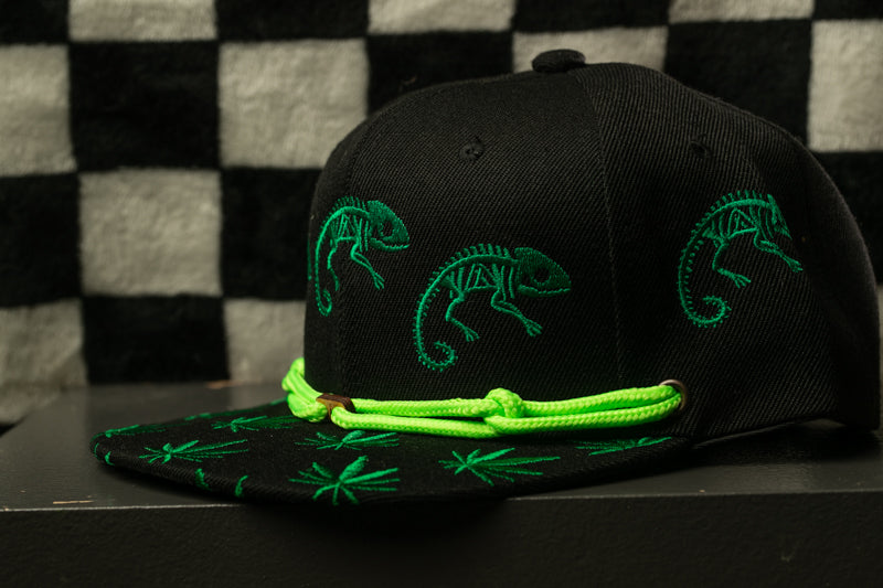 A black baseball cap with green chameleons printed on it, sitting on a black surface against a checkered background.
