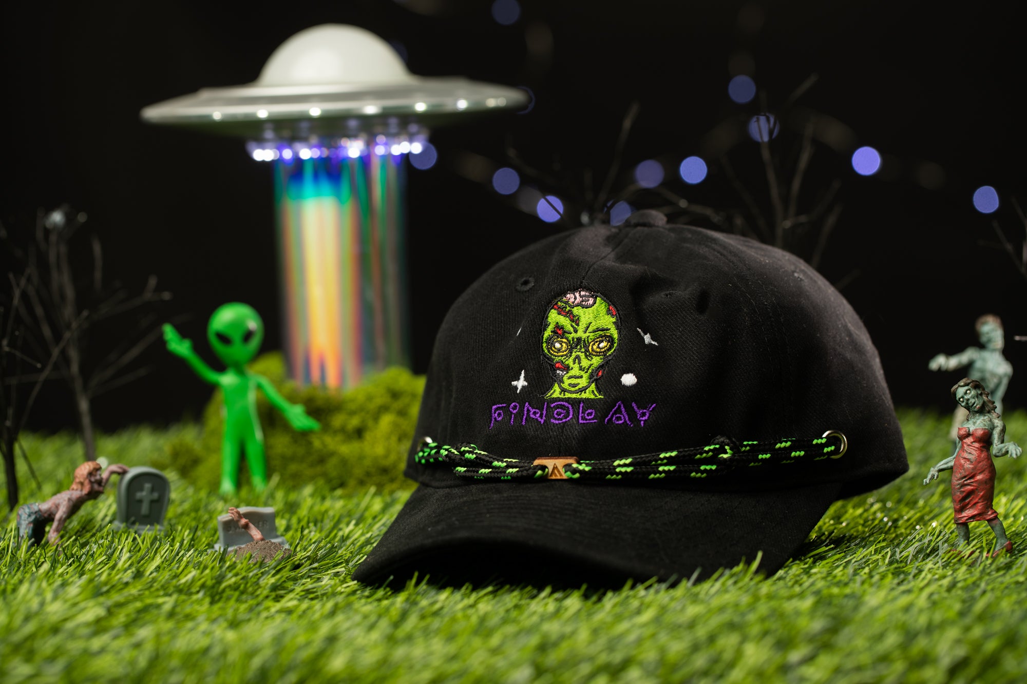 a black baseball cap with a green alien face and the word "PANDEY" printed on it, surrounded by various small toy figures and a green alien figure in the background.