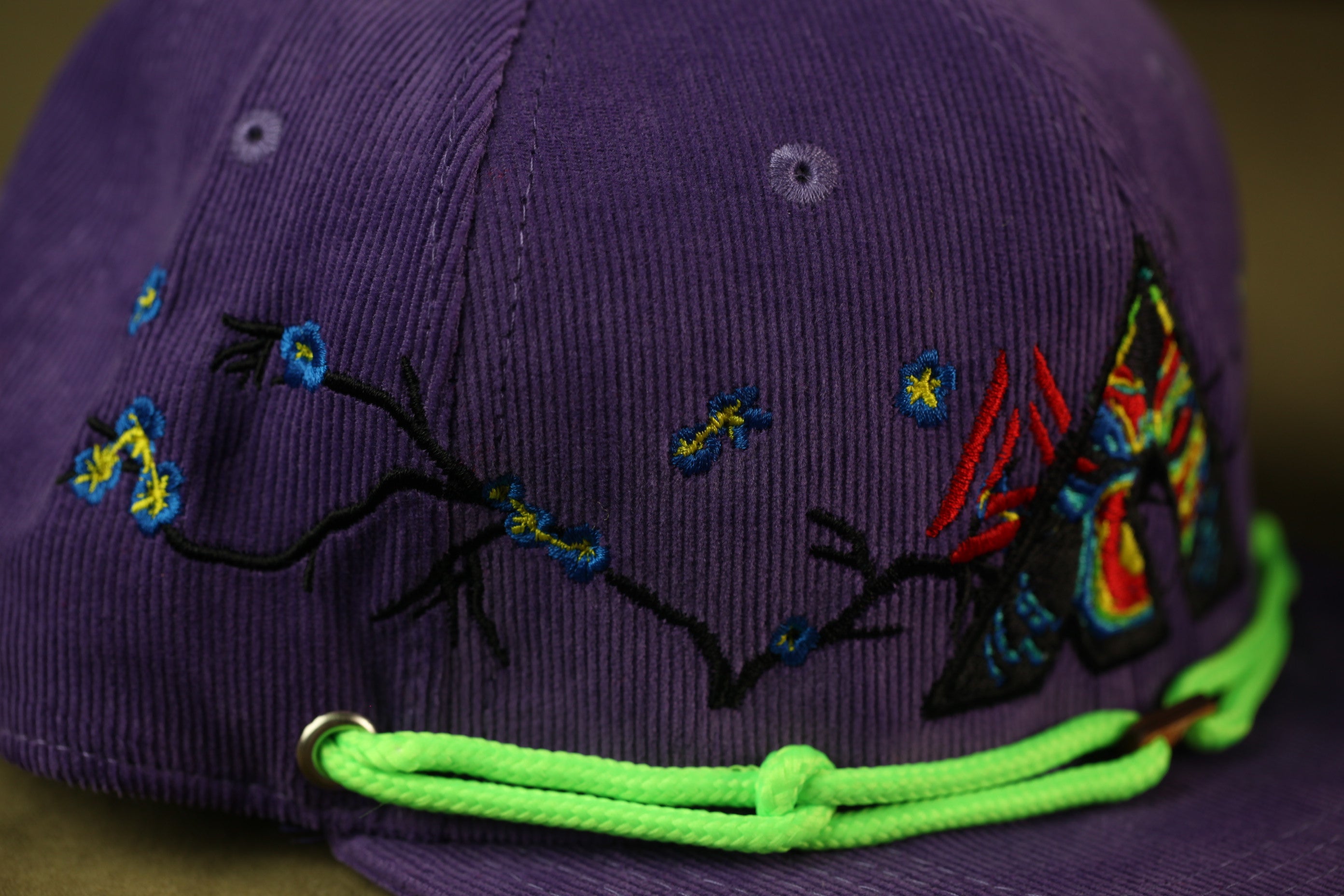 A close-up view of a purple baseball cap with colorful embroidered designs, including flowers and butterflies, and a neon green string.