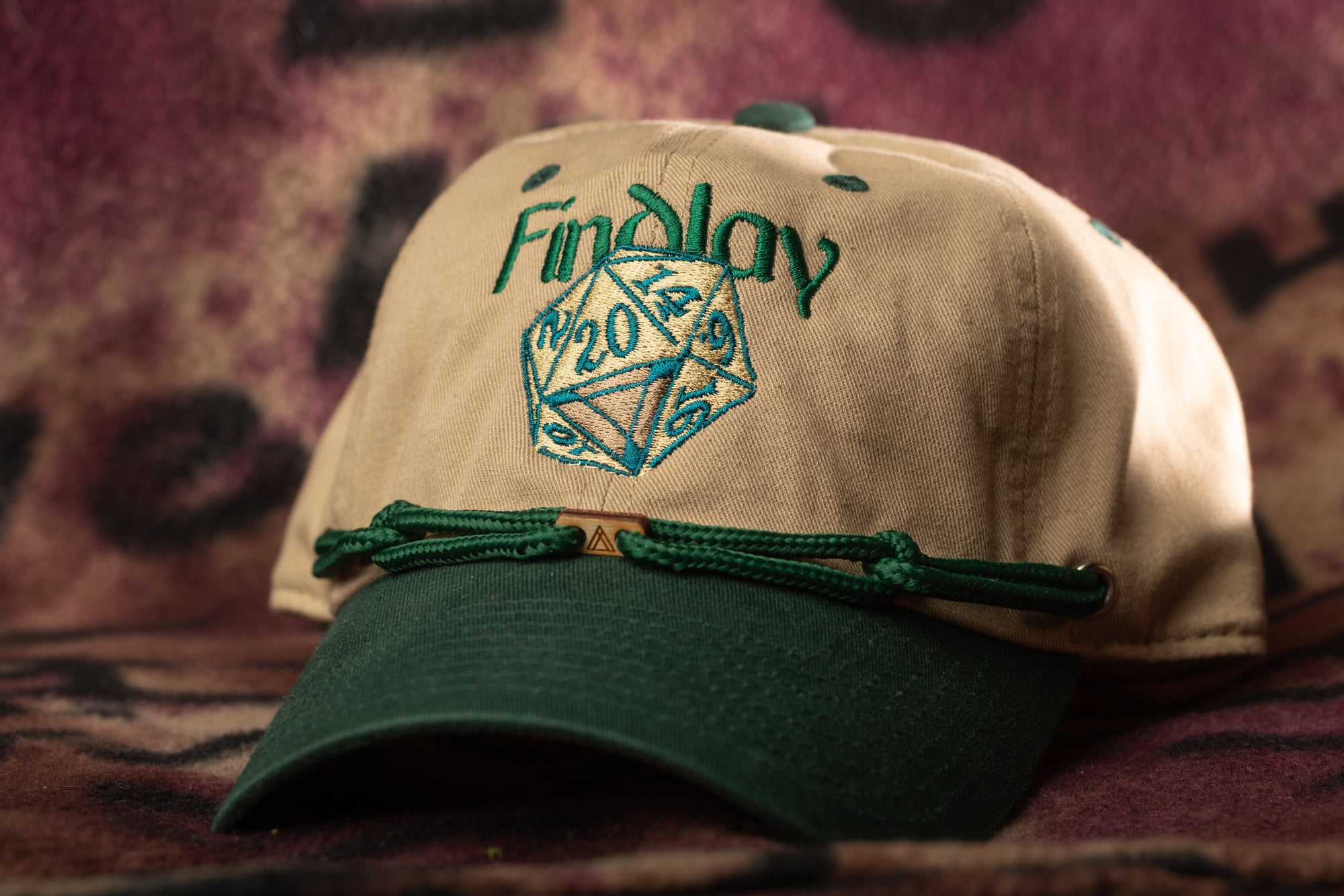 A beige baseball cap with a green brim and a green string, featuring a logo with the text "Findlay" and a dice graphic.
