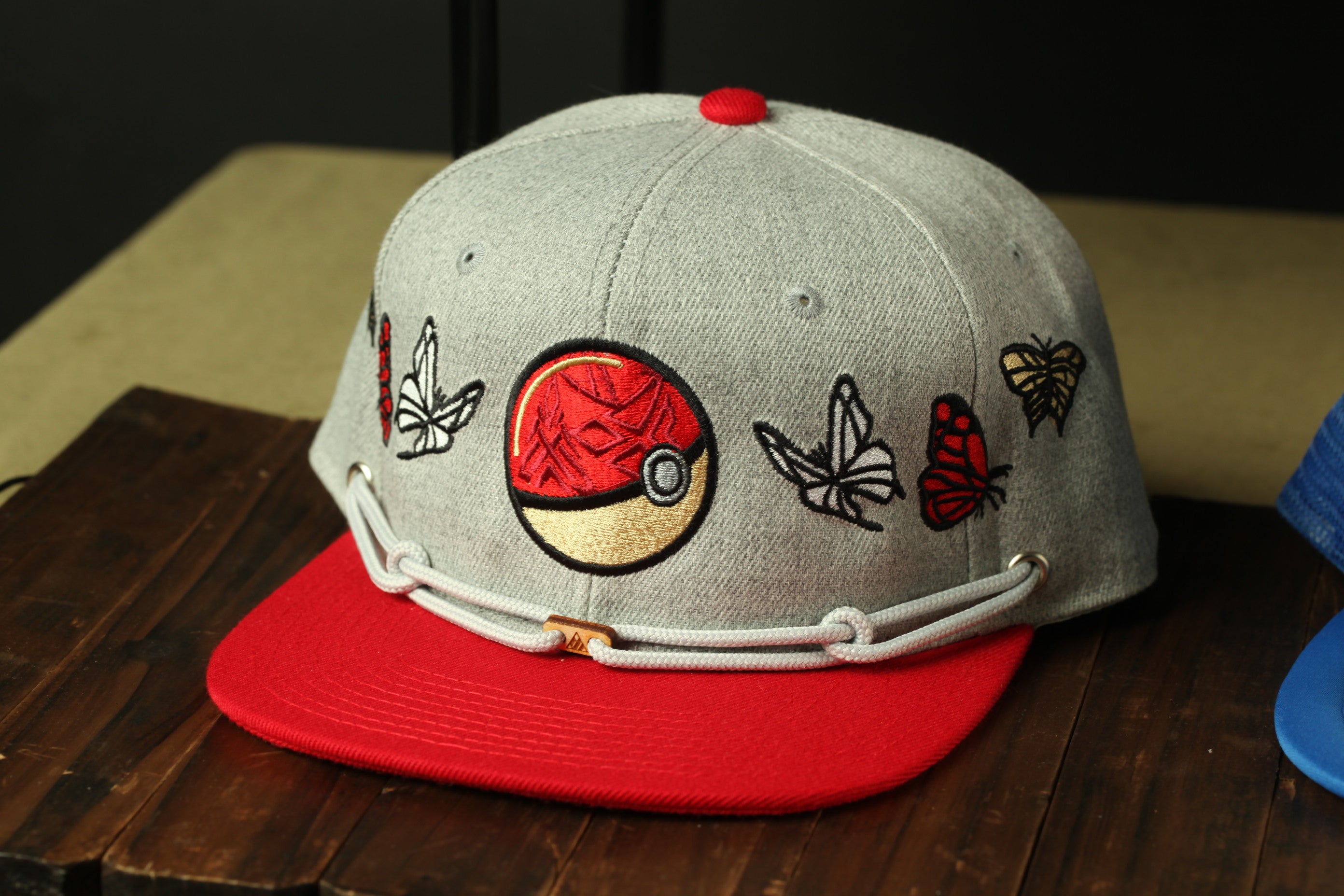 A gray baseball cap with a red brim and a circular logo featuring a Pokemon character, along with various butterfly and flower designs, is displayed on a wooden surface.