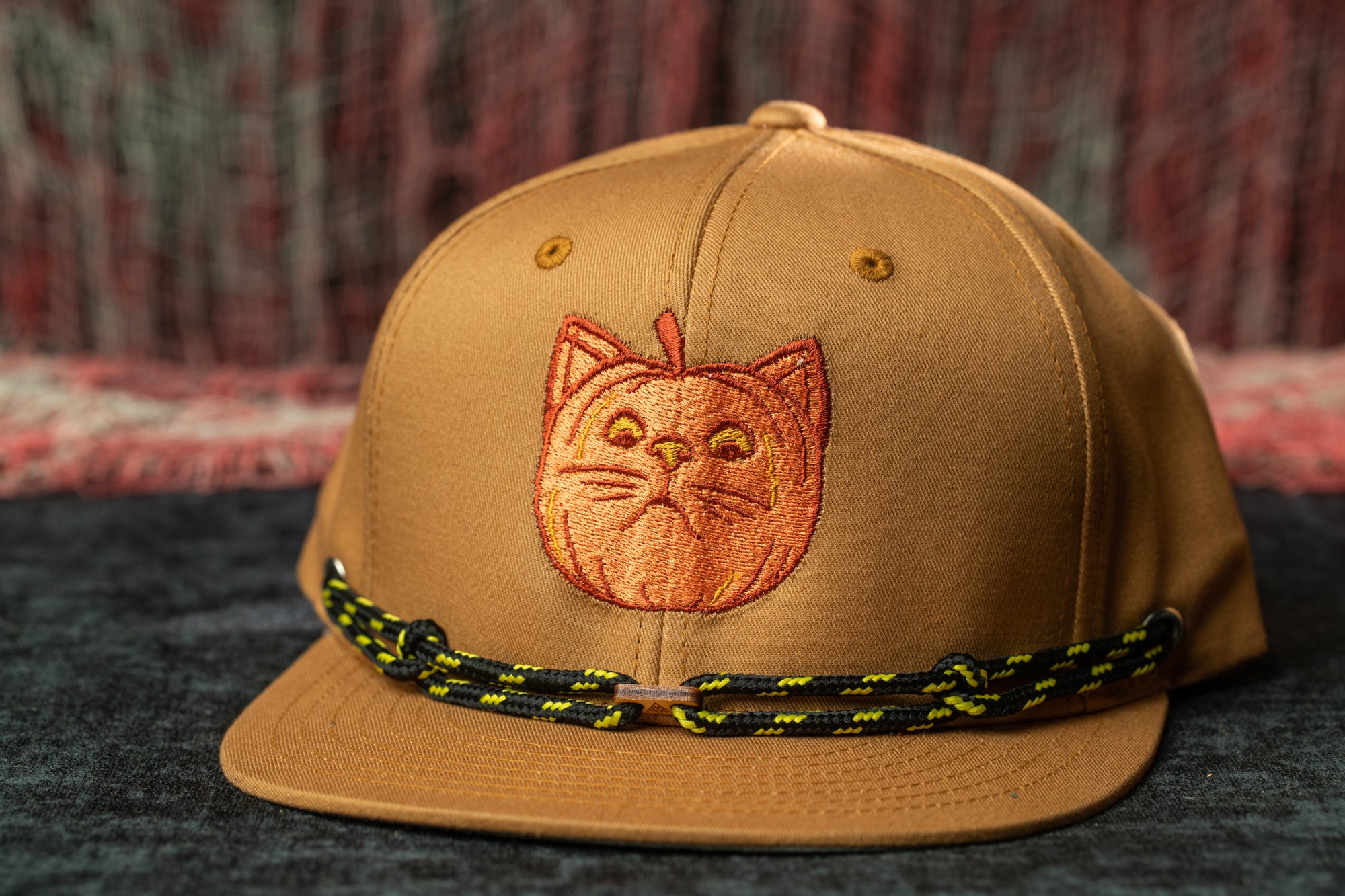A brown baseball cap with a cat embroidered on it, sitting on a dark surface.