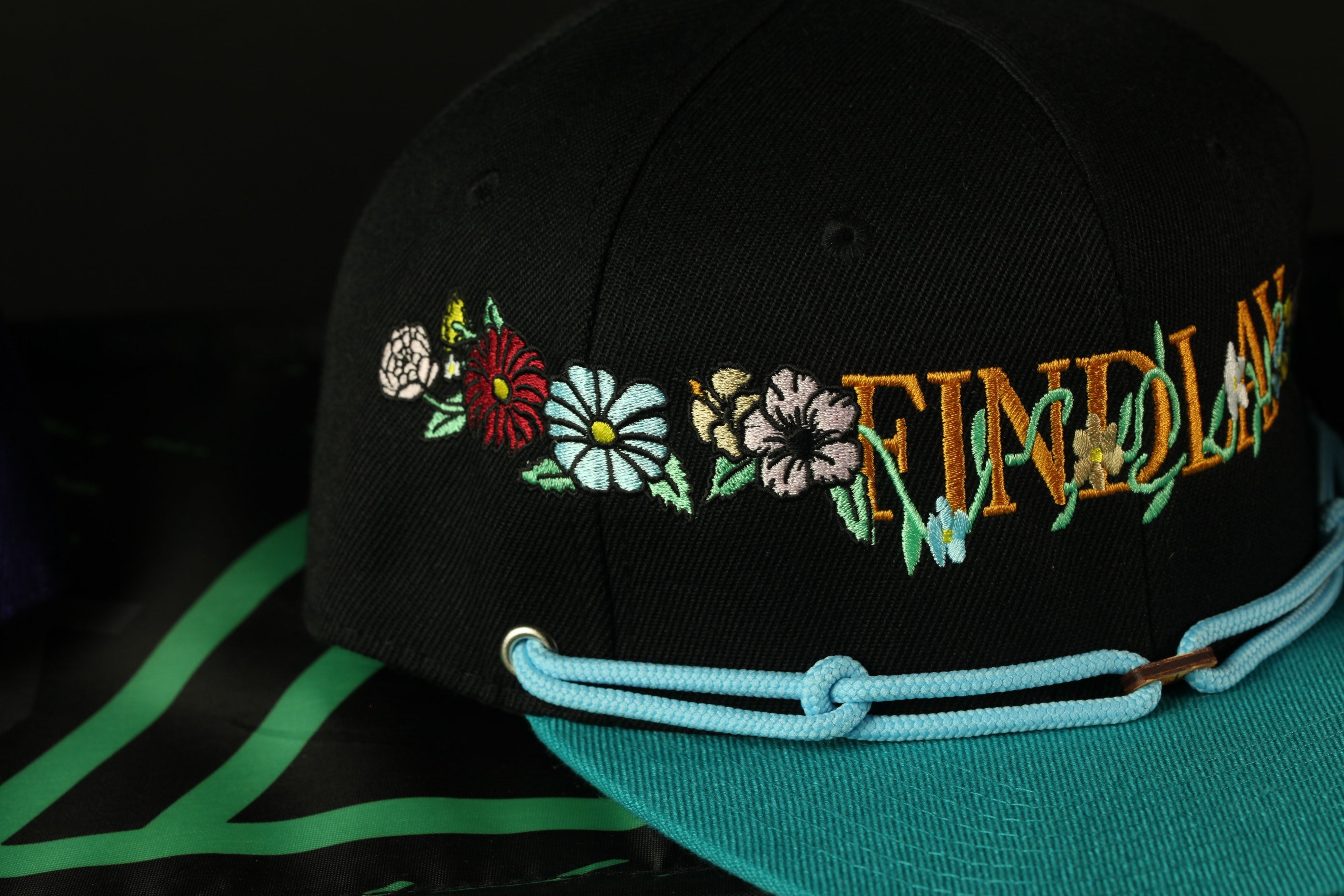 A black baseball cap with colorful floral embroidery and the word "FIND" written on it, placed on a turquoise cap.