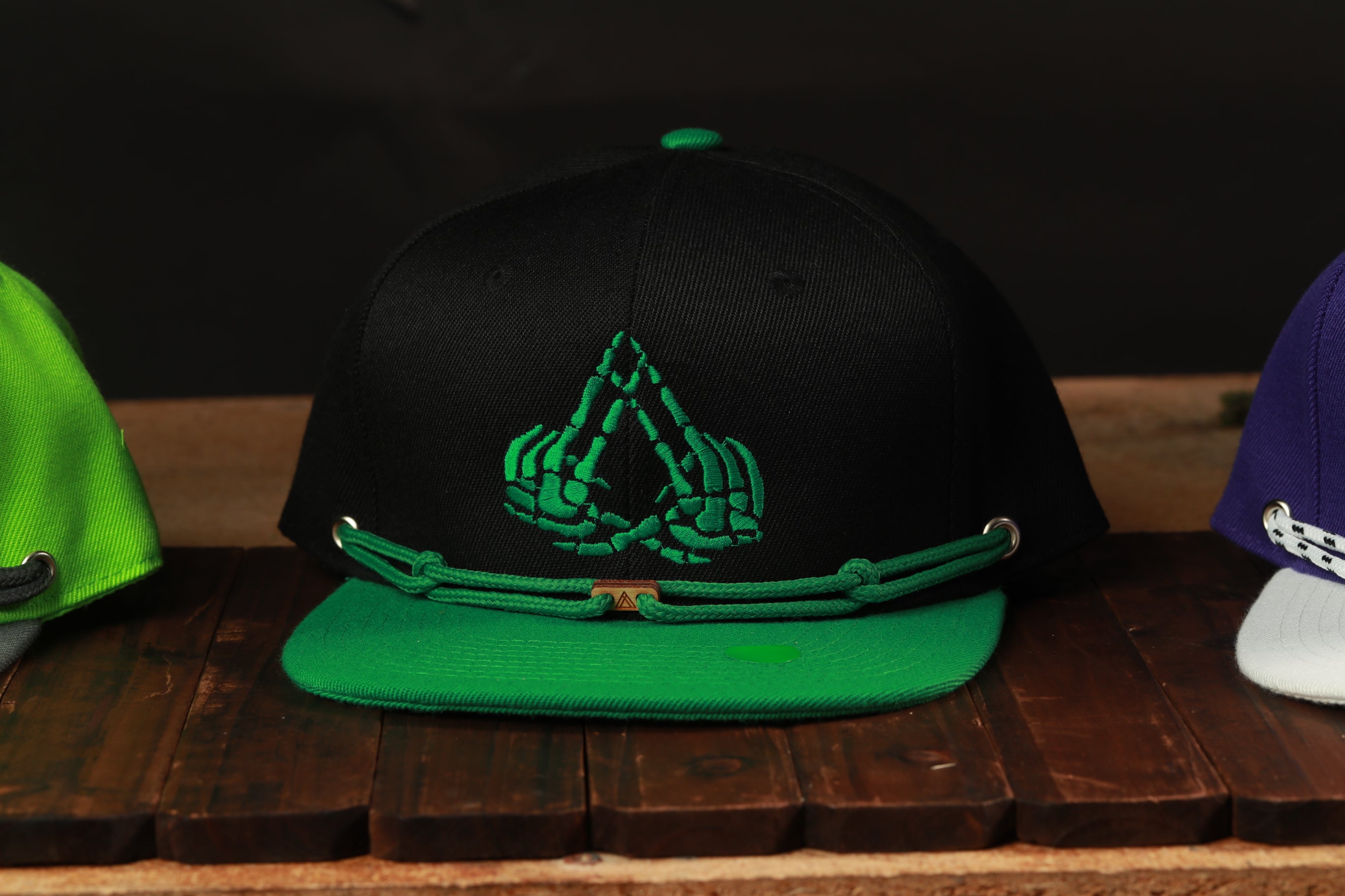A black baseball cap with a green brim and a unique green and black skull-like logo on the front, displayed on a wooden surface.