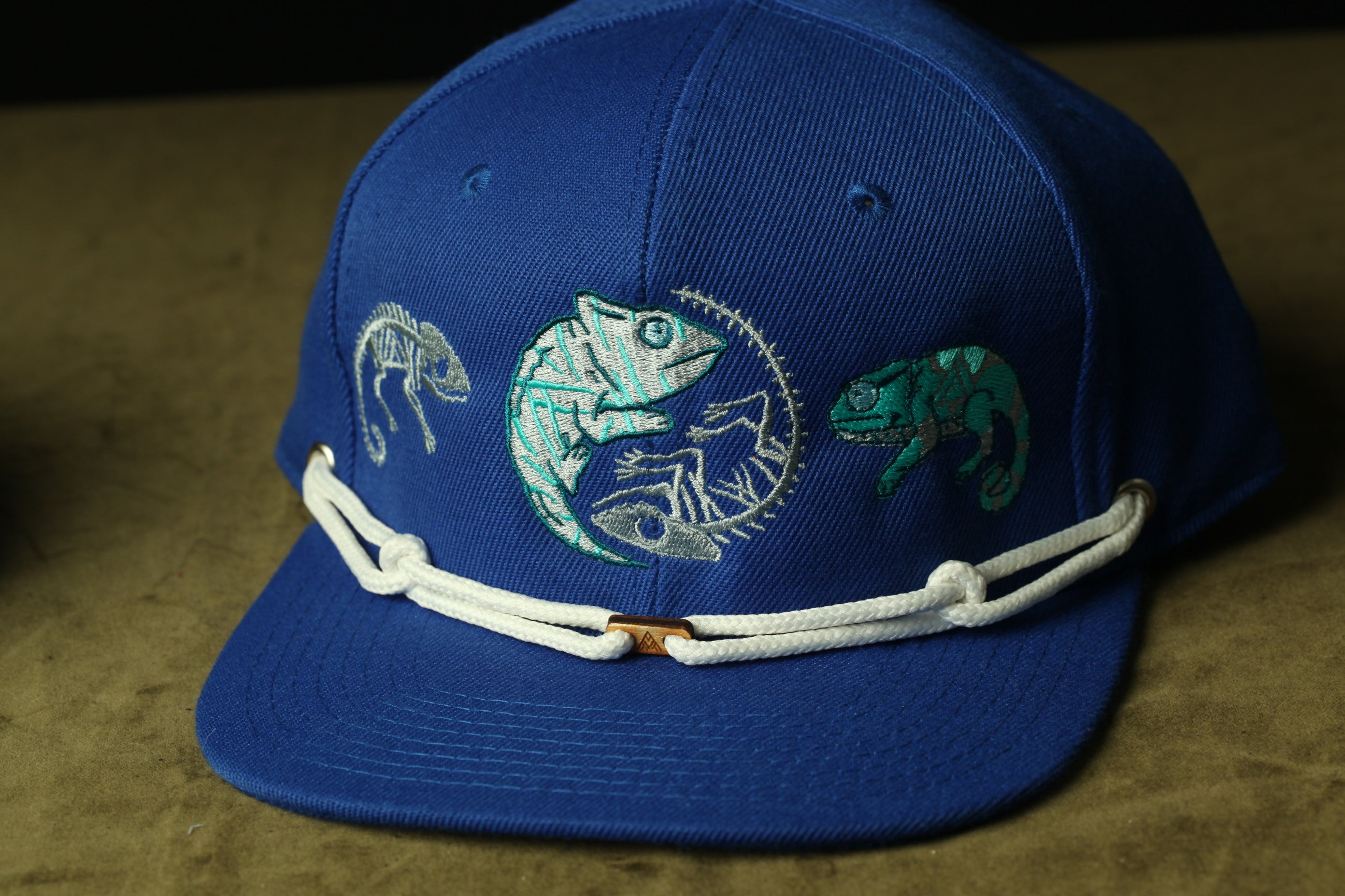 A blue baseball cap with a white lace band and three chameleon designs on the front.