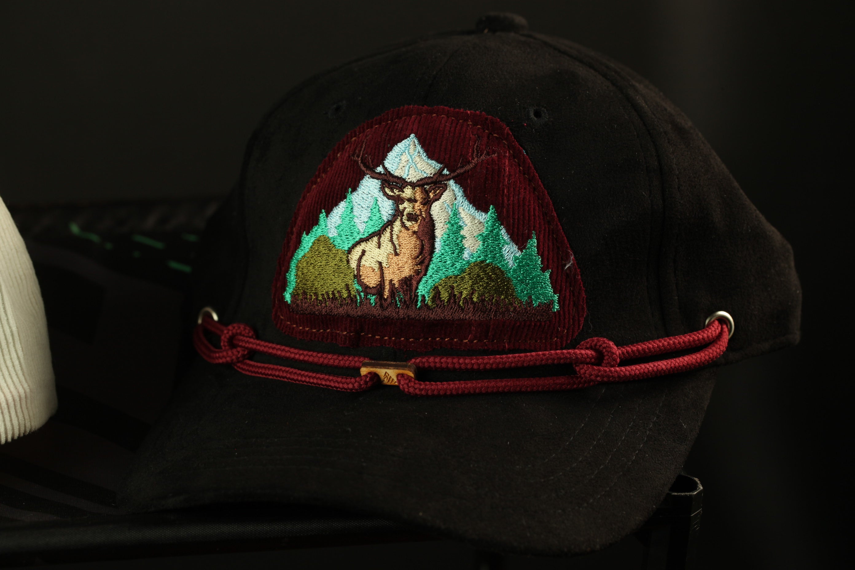 A black baseball cap with a red and green embroidered design depicting a deer standing in a mountainous landscape.