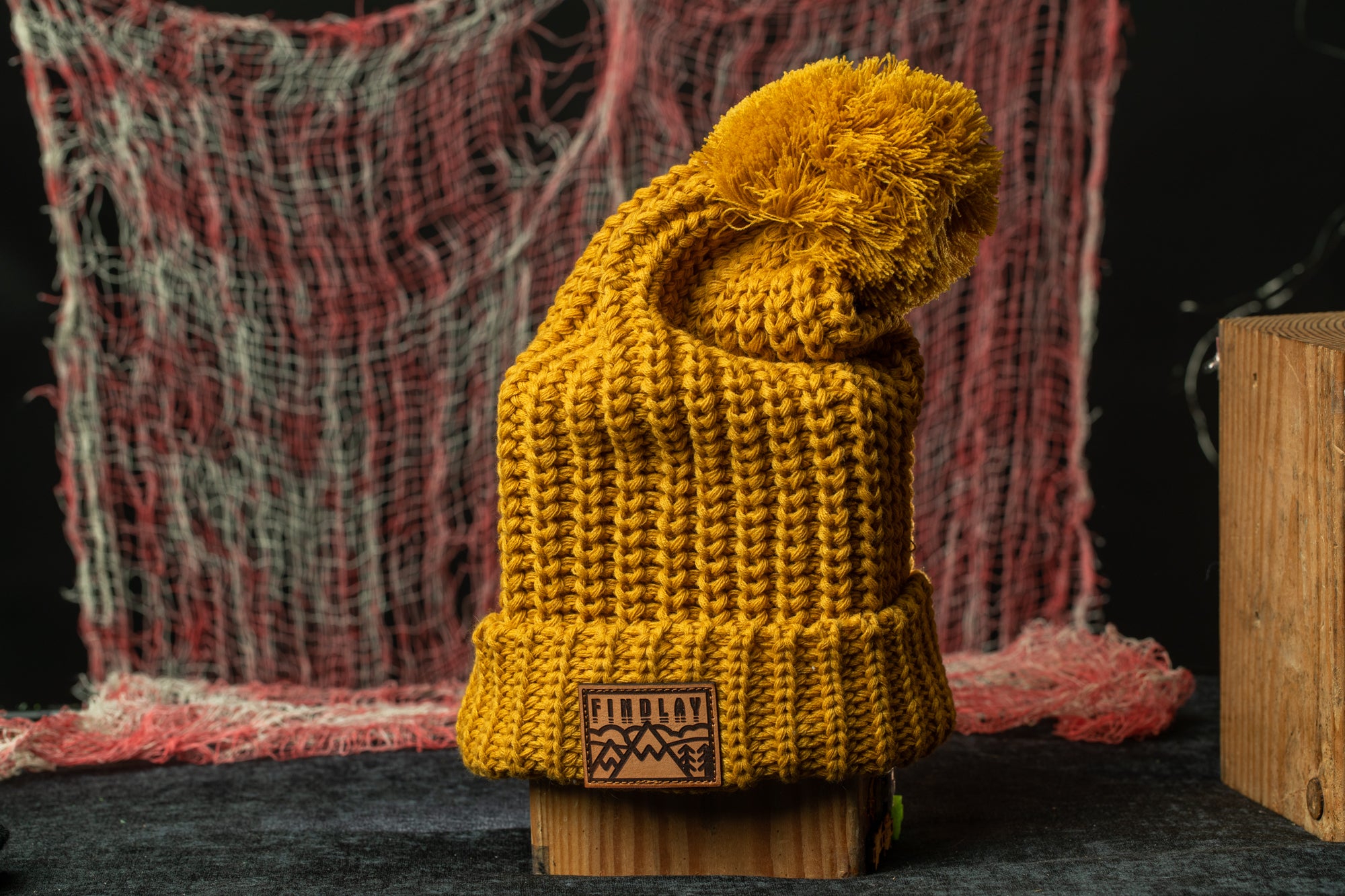 A yellow knit beanie with a pom pom sits on a wooden stand against a red and white background.