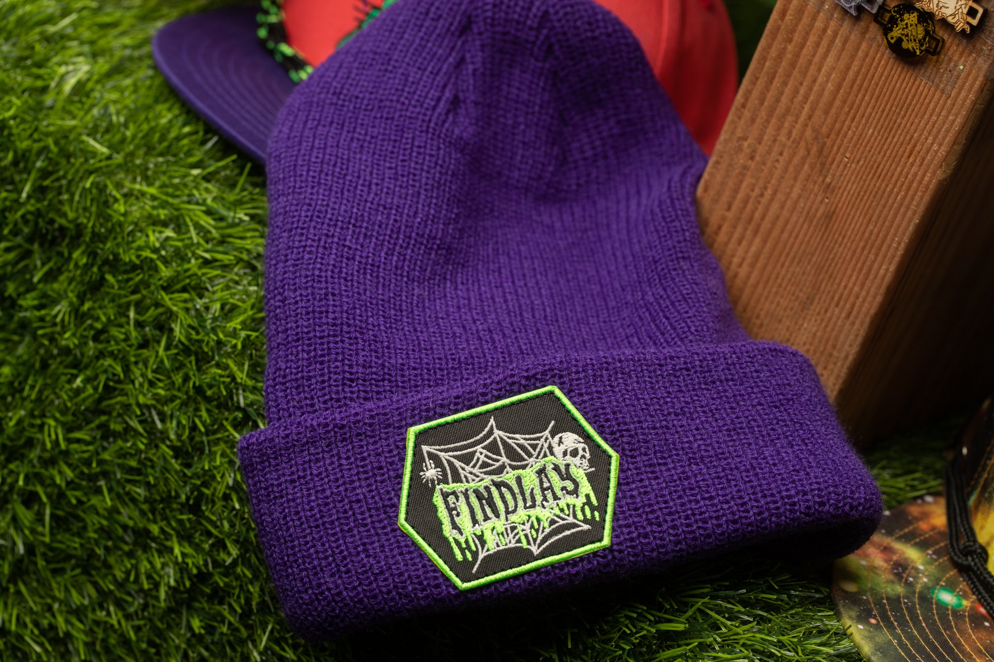 A purple beanie with a green and white spider web design sits on a grassy surface, with a wooden object and a red hat in the background.