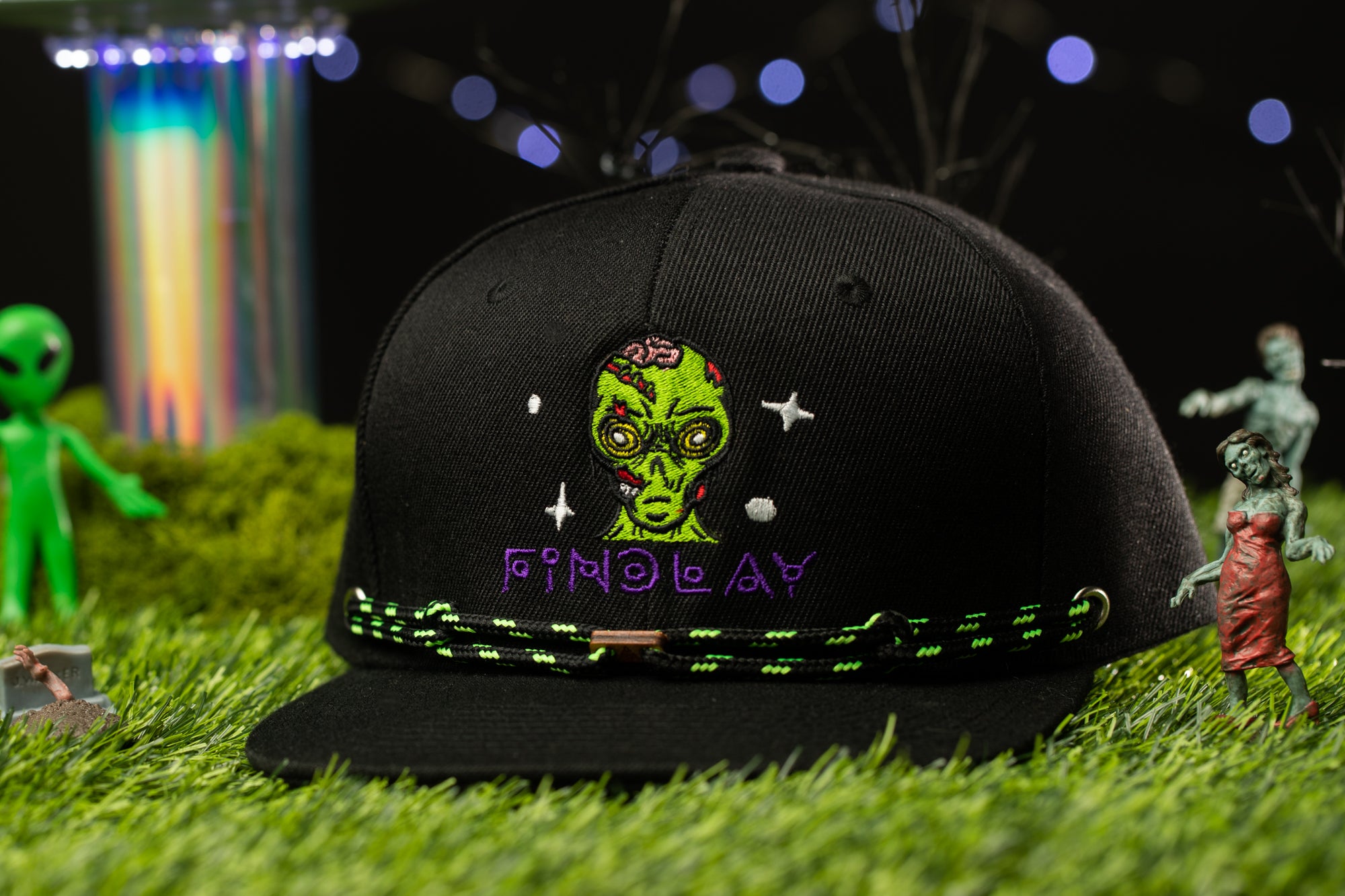 A black baseball cap with a green alien face and purple text is displayed on a grassy surface, surrounded by various small figurines and a colorful, blurred background.