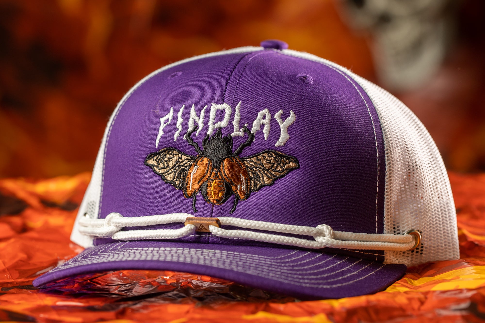 A purple baseball cap with a white mesh panel and a white string attached to the brim, featuring an embroidered design of a bee on the front.