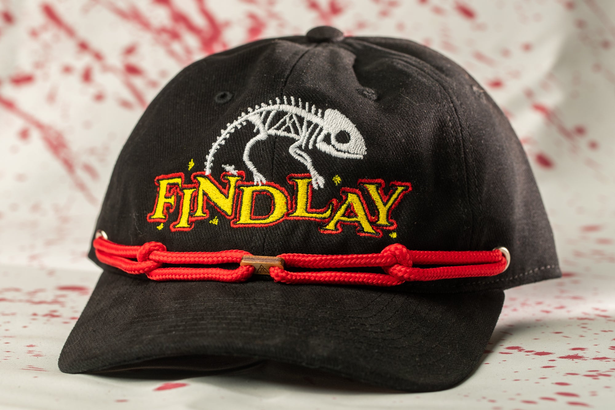 A black baseball cap with a red string and a skeleton design on it, featuring the word "FINDlay" in yellow letters.