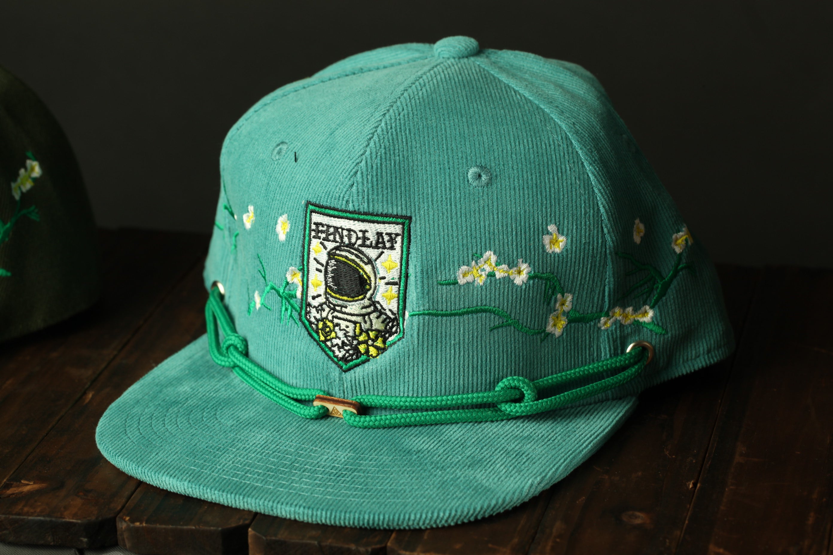 A turquoise baseball cap with a floral embroidered design and a small patch on the front, sitting on a wooden surface.