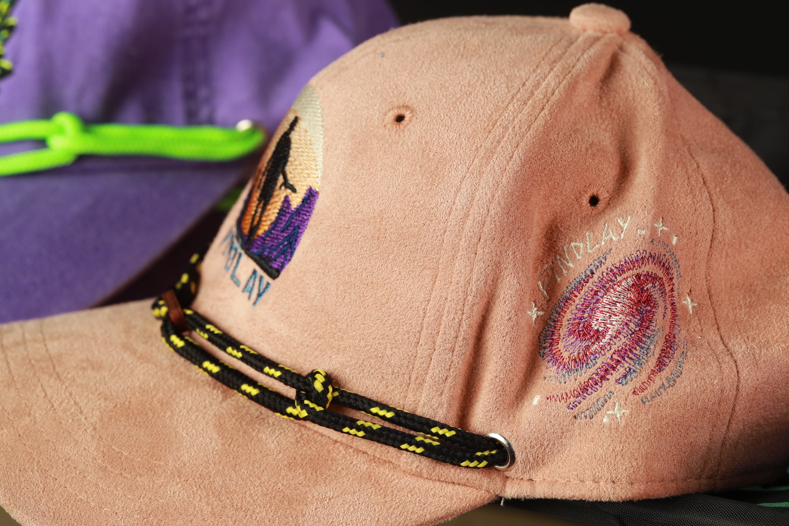 A close-up image of a pink baseball cap with embroidered designs and a black and yellow string.