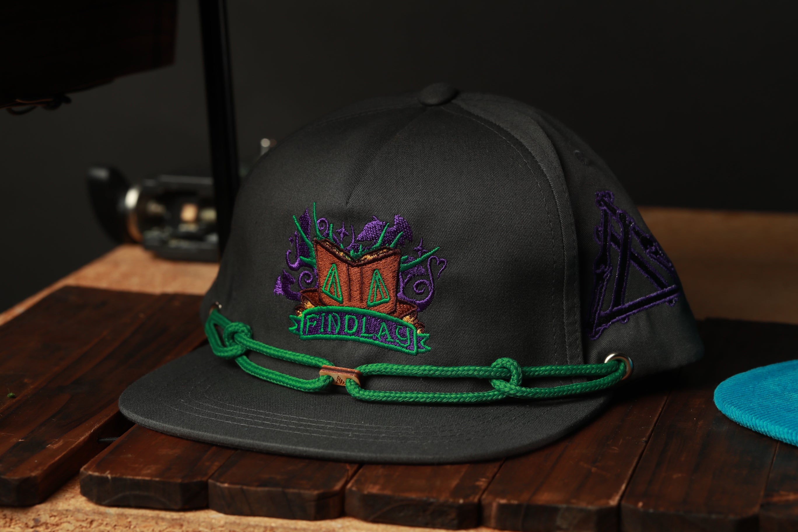 A black baseball cap with a green and purple embroidered logo, sitting on a wooden surface.