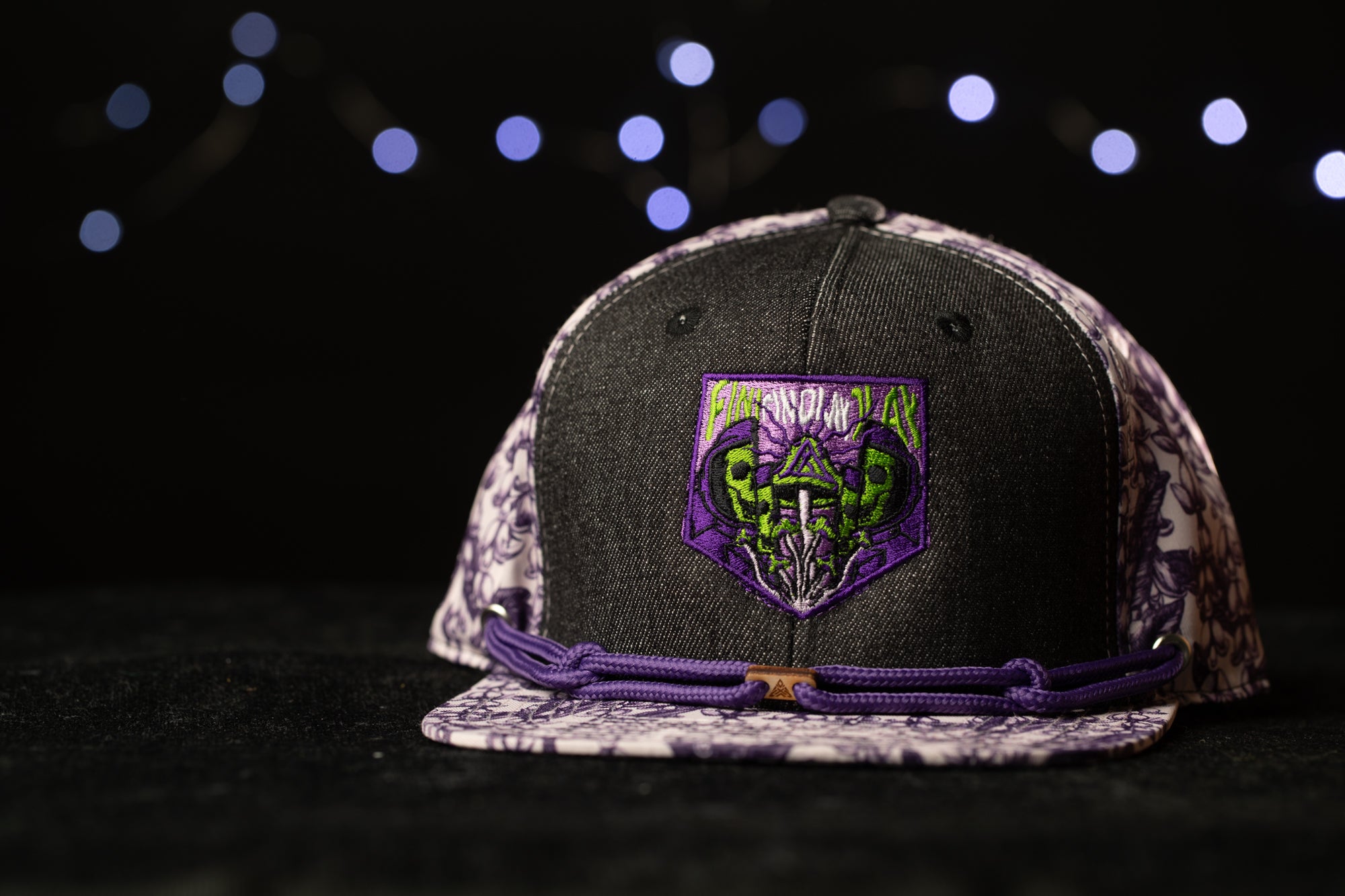 A black baseball cap with a purple and white floral pattern and a green and purple design on the front, placed on a dark surface against a background of blurred lights.