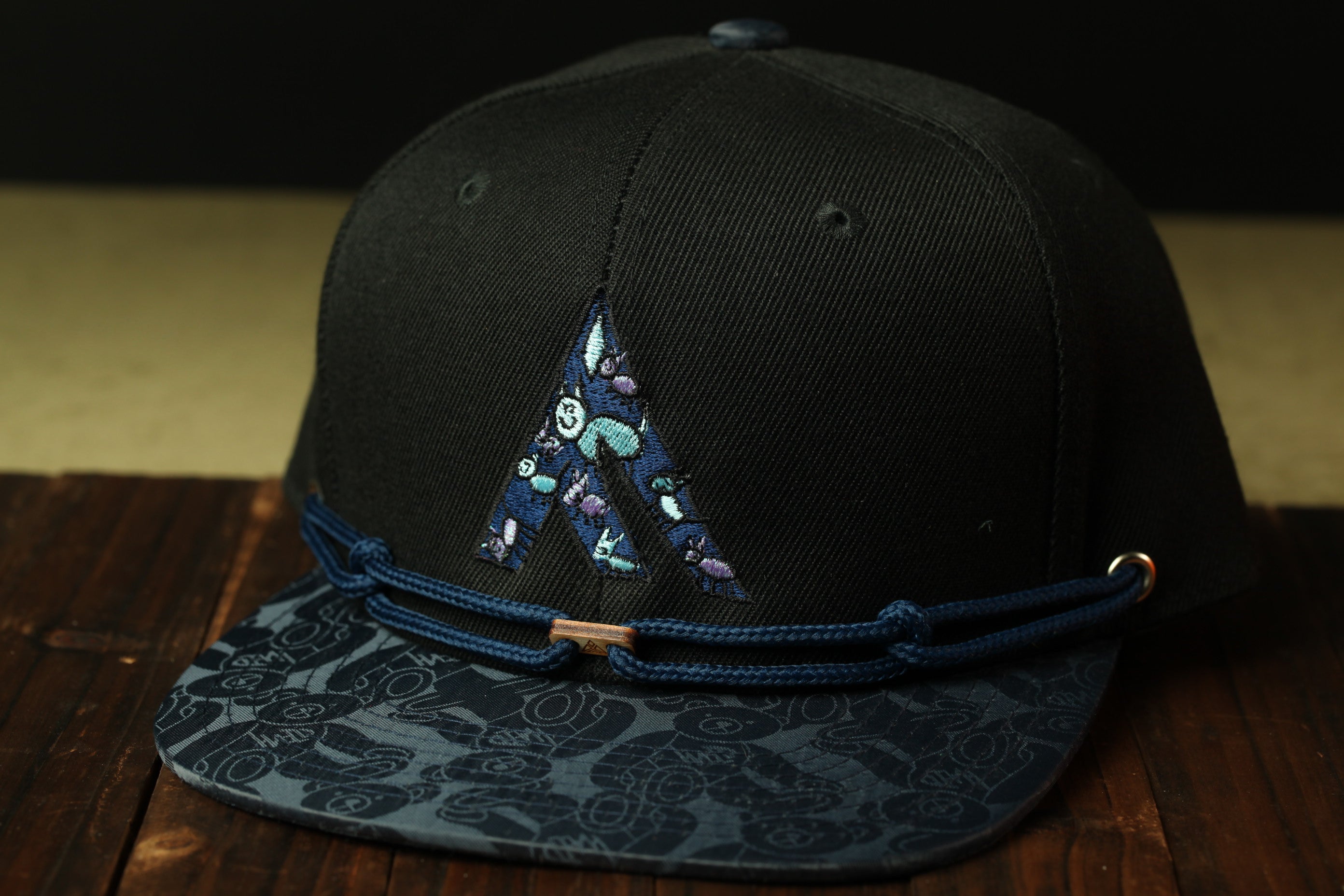 A black baseball cap with a blue and black patterned brim and a blue string around the brim, placed on a wooden surface.