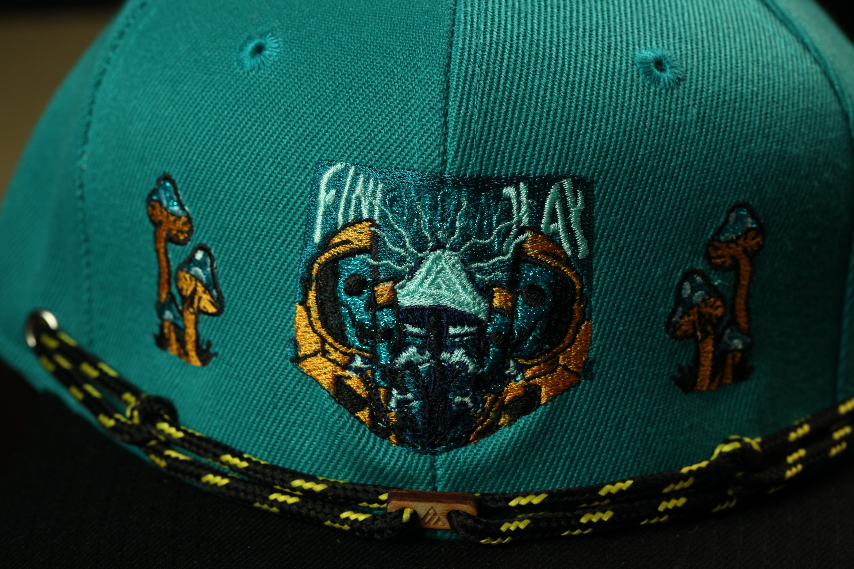 A close-up view of a teal-colored baseball cap with a unique embroidered design featuring a bear's face and mushrooms.