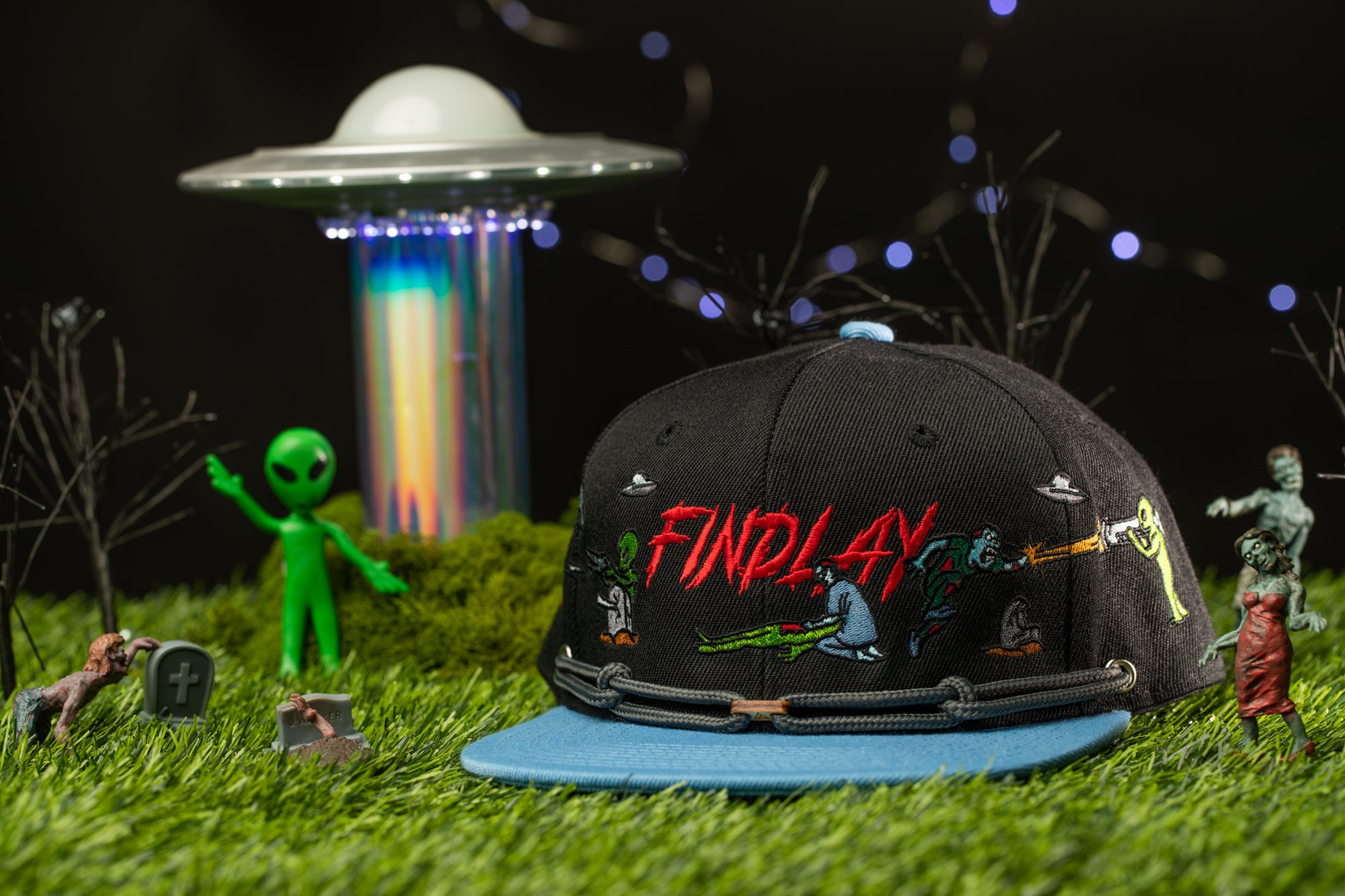A black baseball cap with the word "FINDLAY" printed on it, surrounded by various toy figures and a green alien figure, all set against a dark background with a large alien spacecraft in the background.