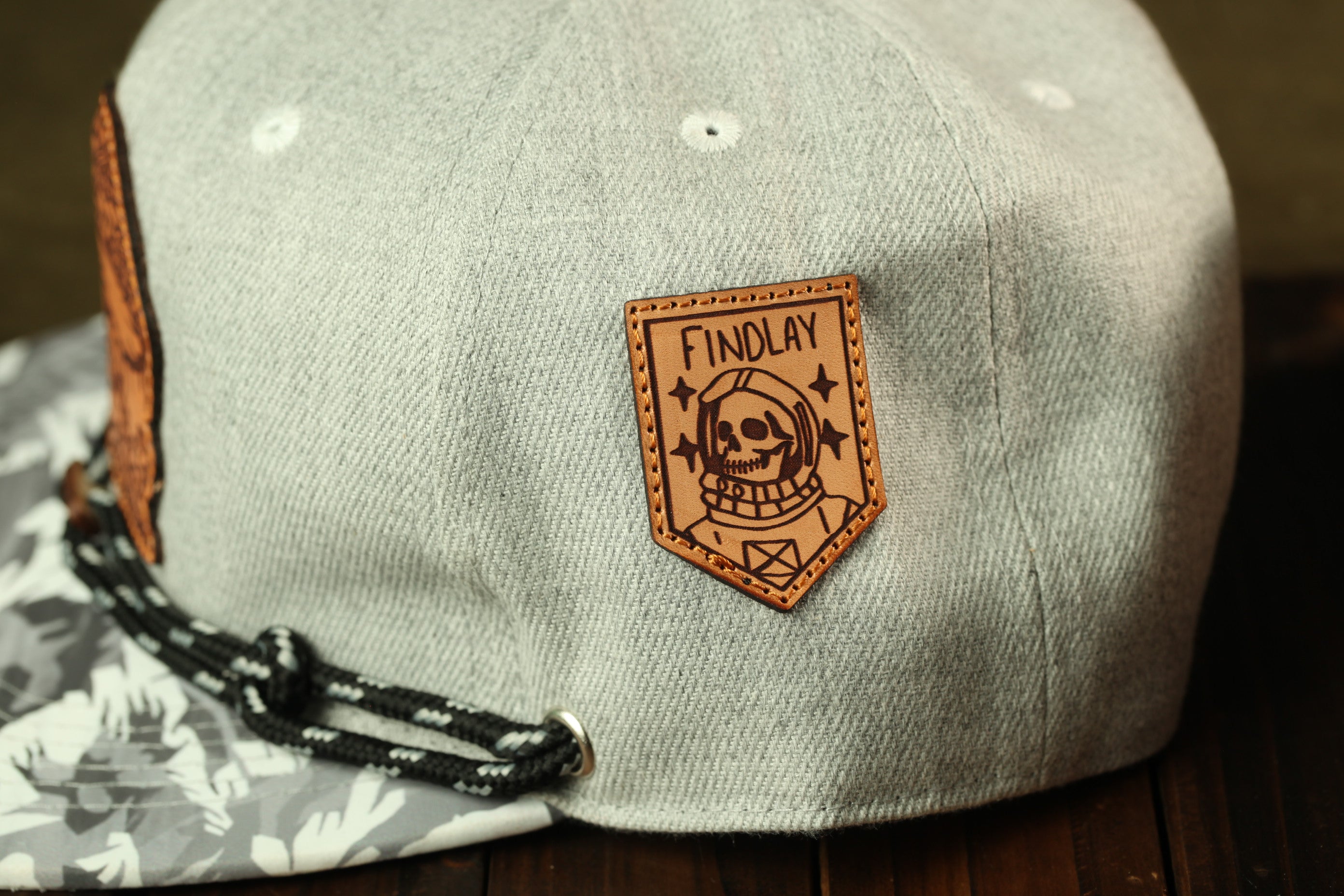 A gray baseball cap with a leather patch on the front, featuring a skull and the text "FINDLAY" printed on it.