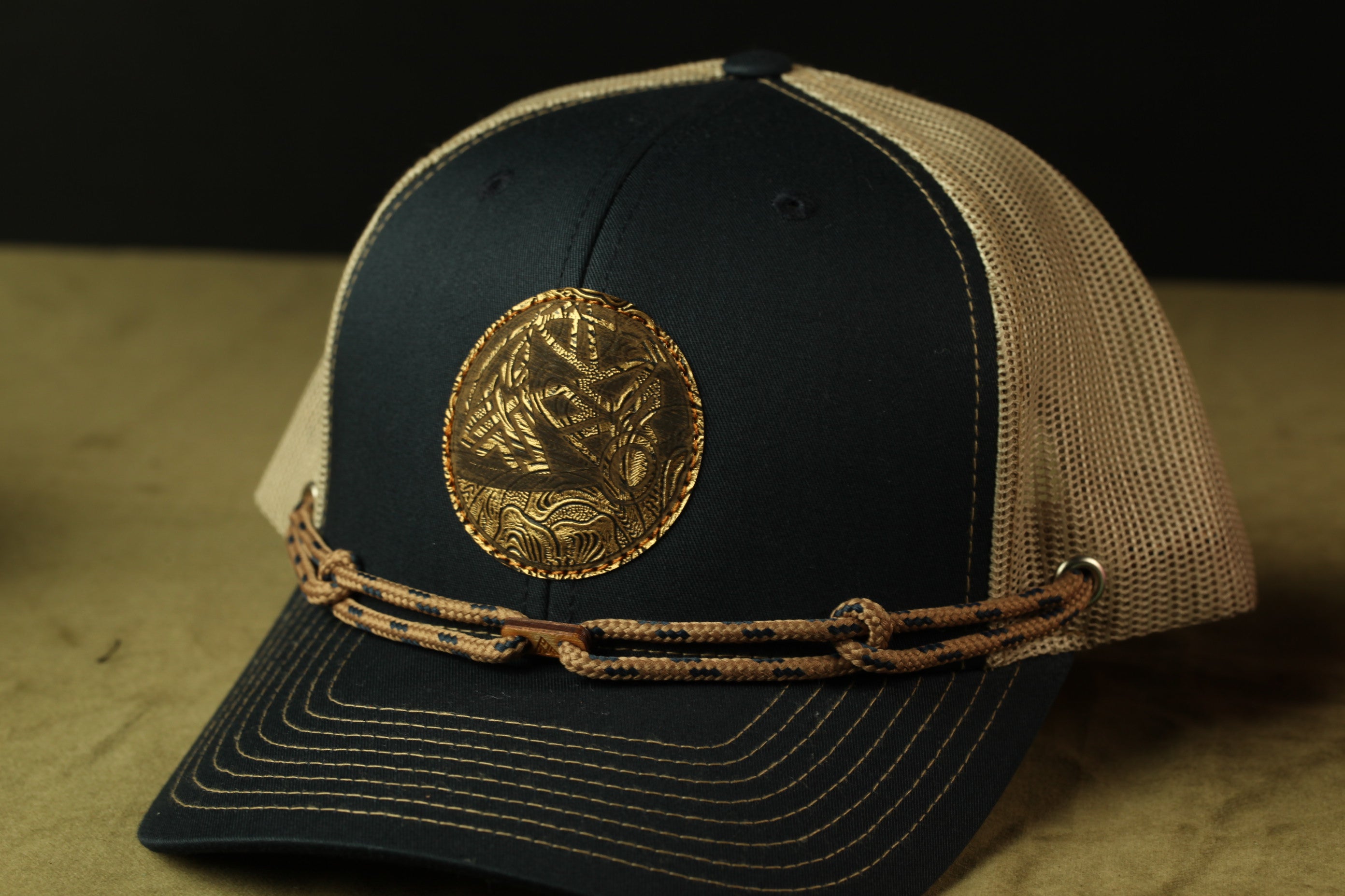 A black baseball cap with a gold emblem on the front, and a beige mesh panel on the side.
