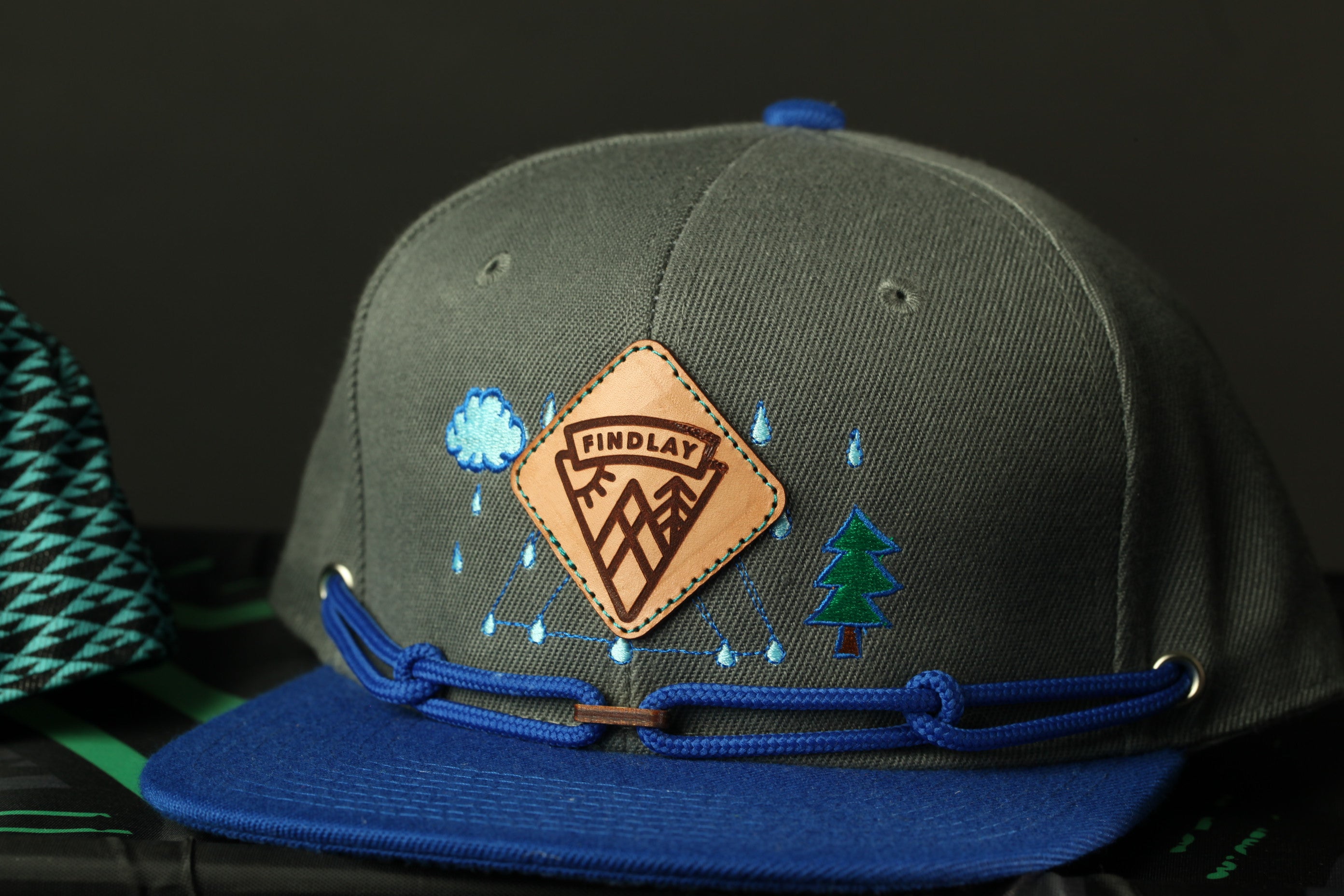 A gray baseball cap with a blue brim and a leather patch featuring a mountain and raindrops, along with the text "FINDA" and a tree design.