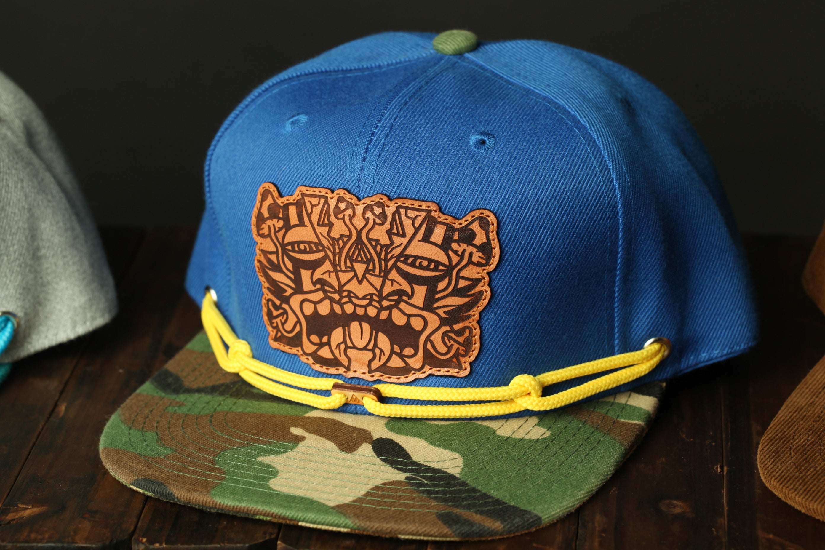 A blue baseball cap with a camouflage pattern and a leather patch featuring an abstract design is displayed on a wooden surface.