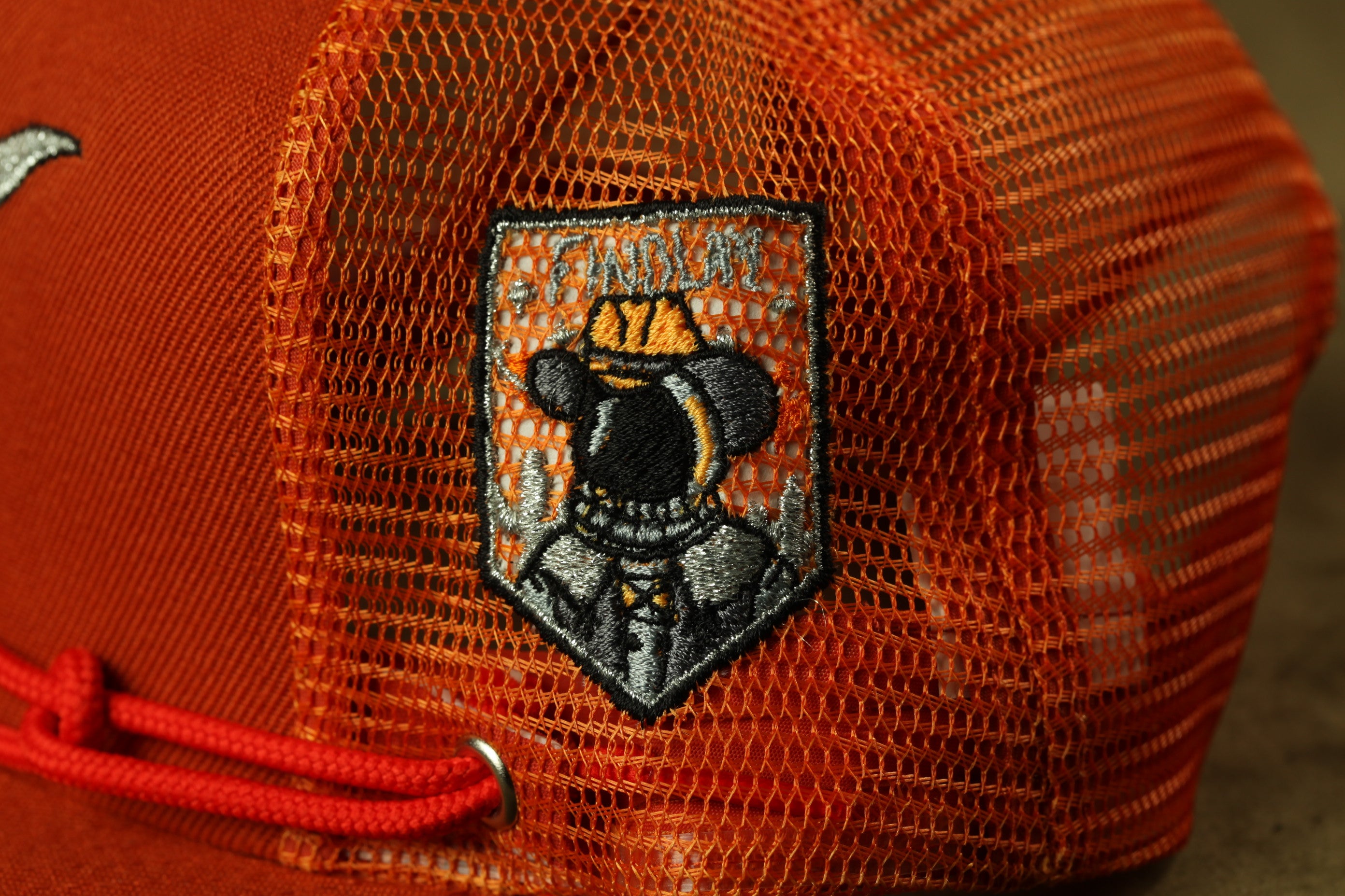 a close-up view of a red mesh hat with a small patch featuring a black and orange design, including a figure wearing a hat and a coat.