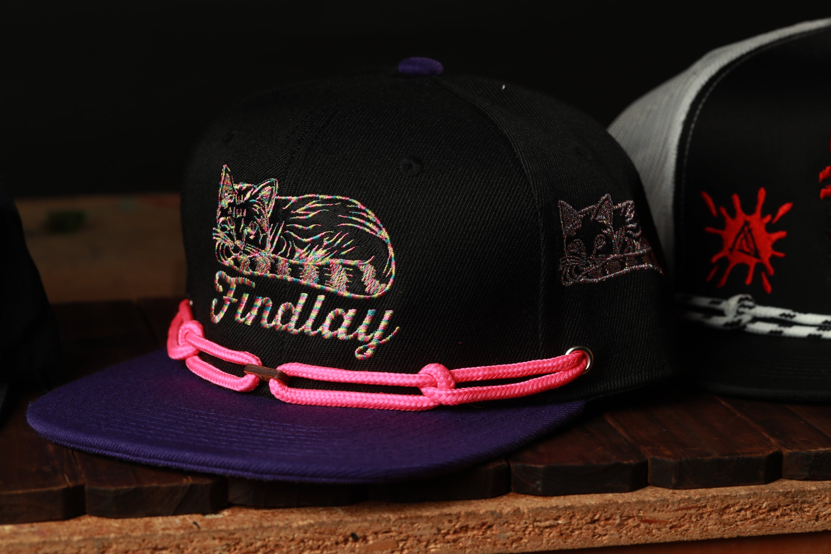 A black baseball cap with a pink chain strap and a cat embroidered on it, sitting on a wooden surface.