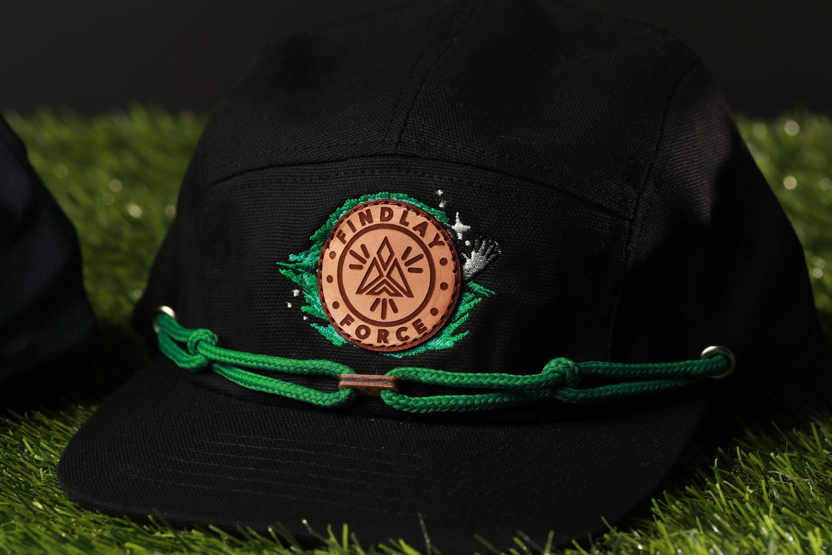 A black baseball cap with a green band and a circular logo on the front, placed on a grassy surface.