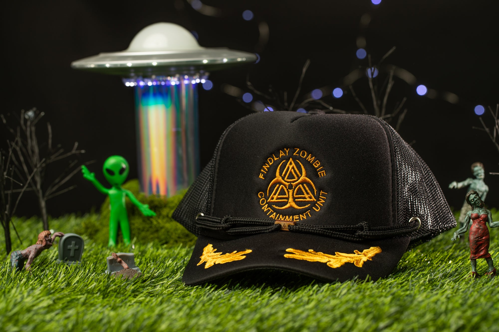 a black baseball cap with gold embroidery, surrounded by various toys and figurines, including a green alien figure, a zombie, and a spaceship.