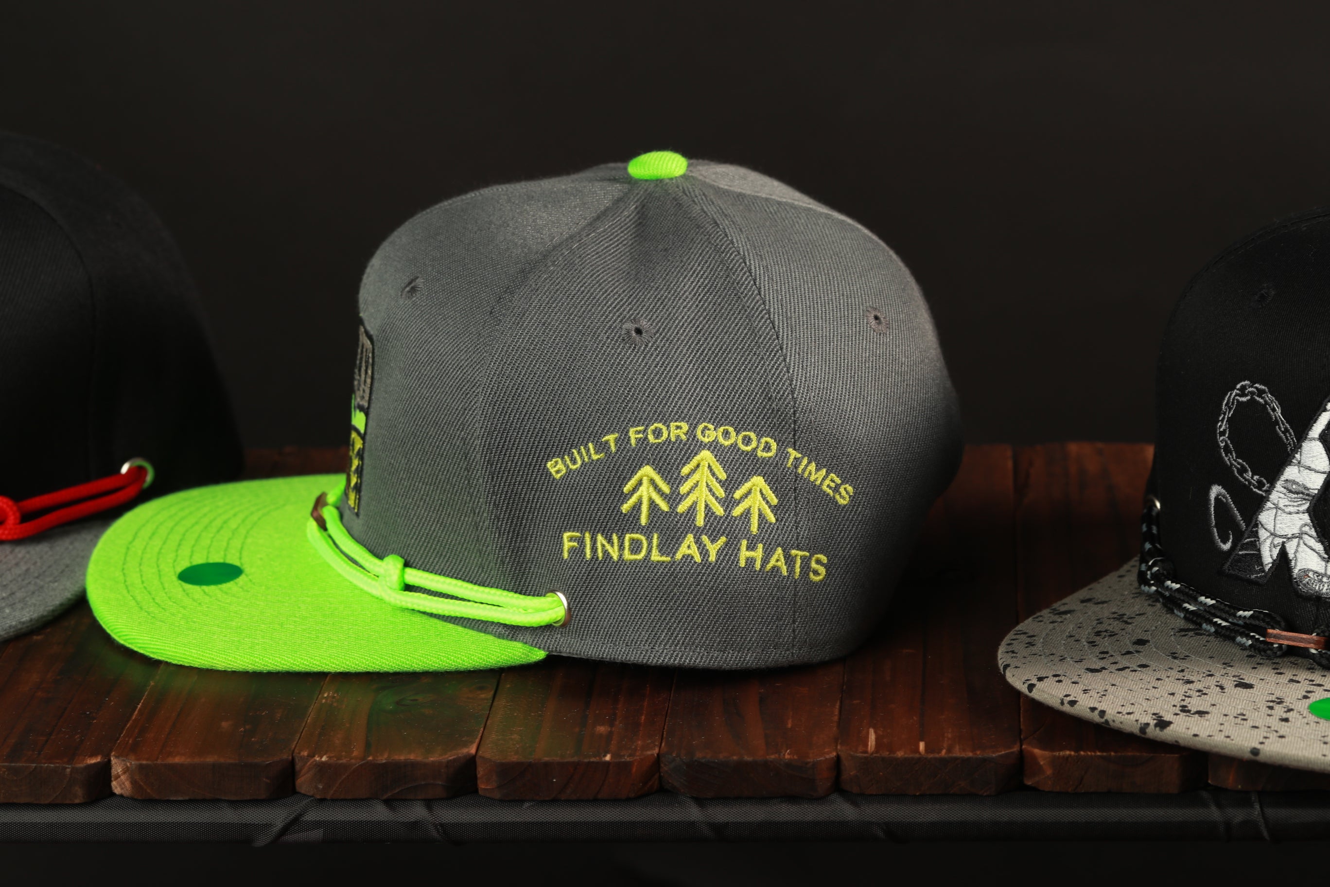 three baseball caps on a wooden surface, with one gray cap with a green brim and yellow text, and two other caps in different colors.