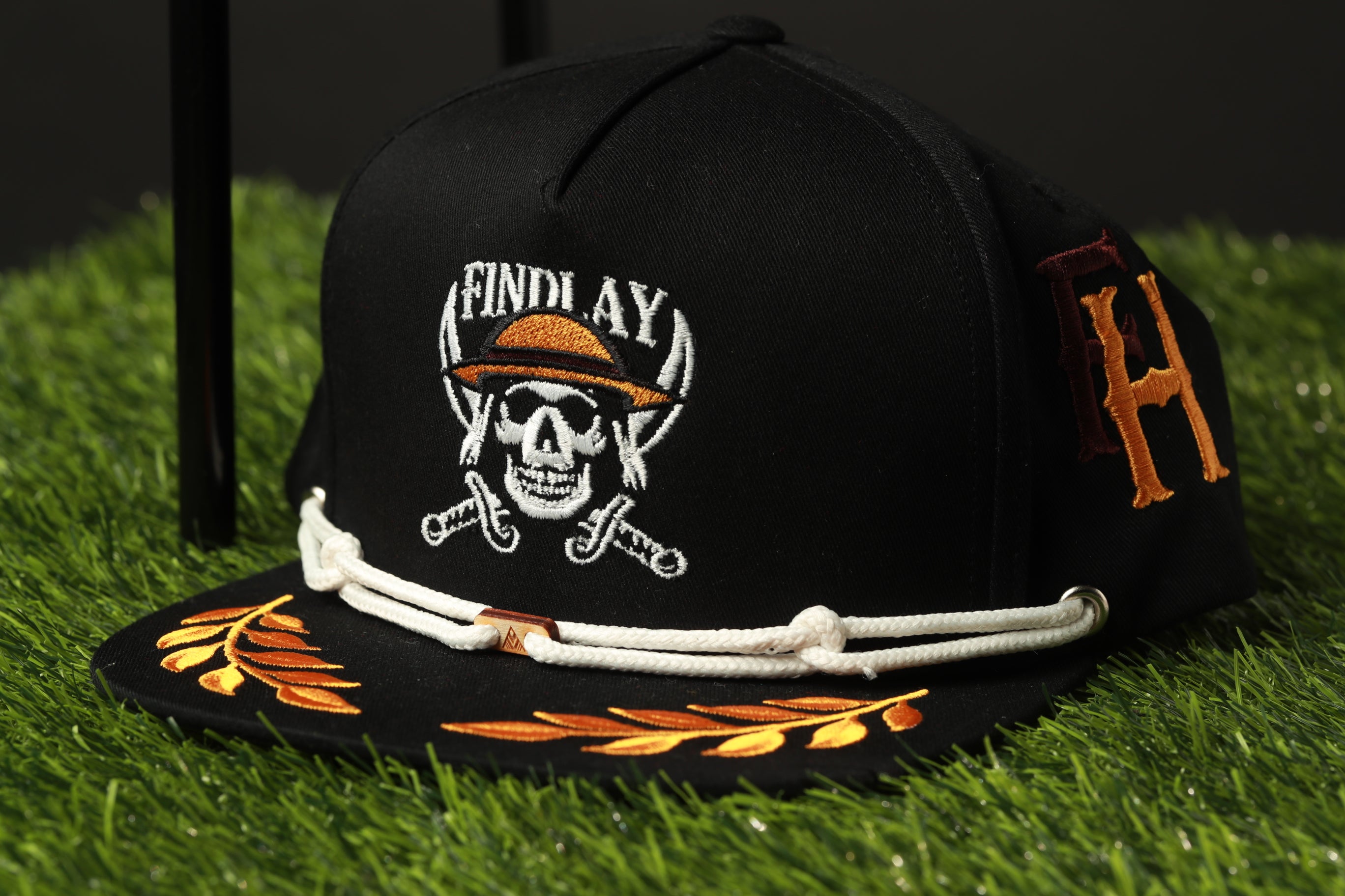 A black baseball cap with a skull and crossbones design, a gold laurel leaf pattern, and a white rope trim, placed on a grassy surface.
