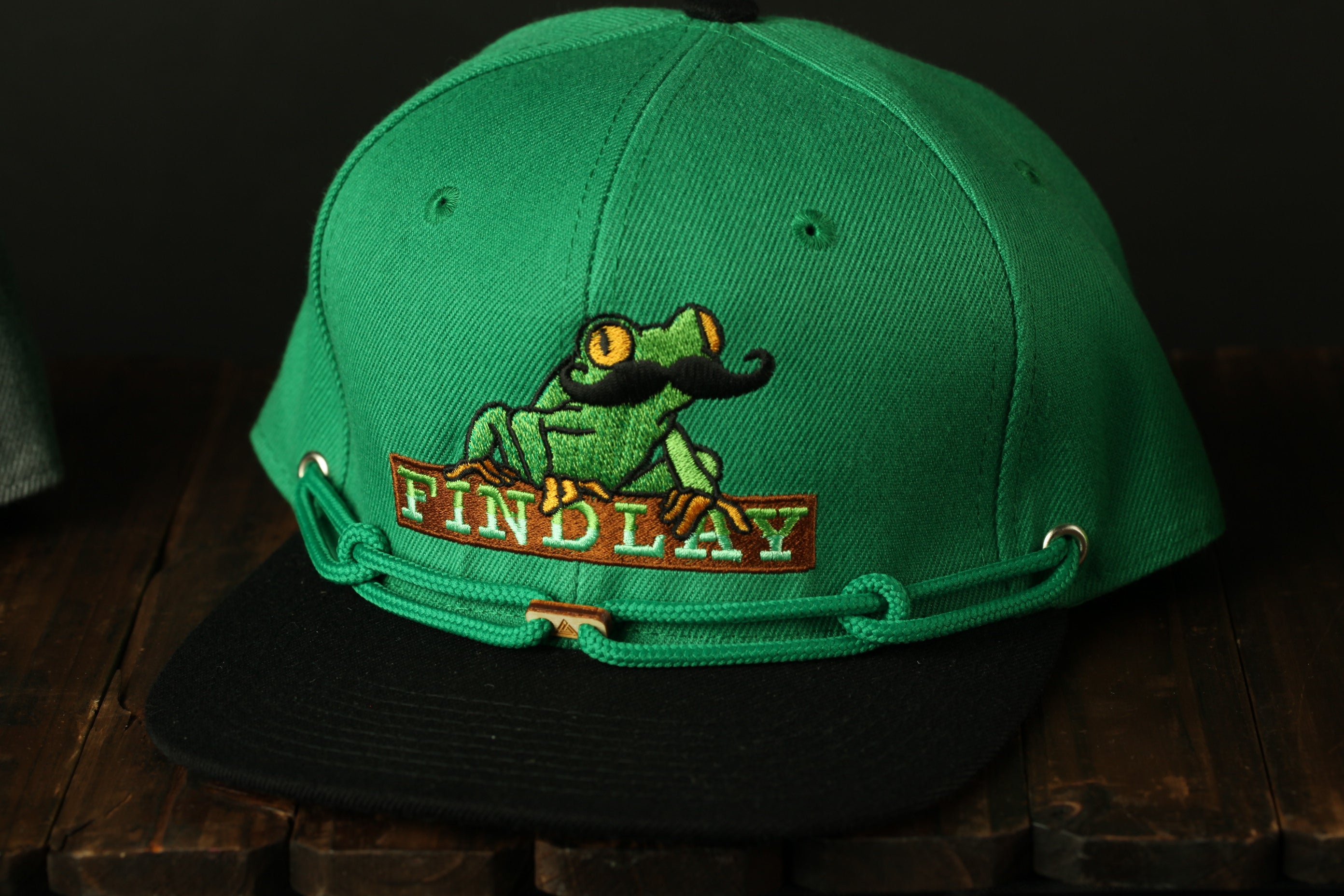 A green baseball cap with a frog embroidered on it, sitting on a wooden surface.