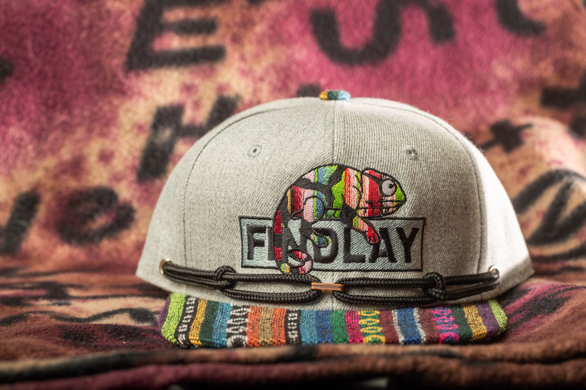 A gray baseball cap with a colorful, embroidered design and the word "FIDDLY" printed on it, placed on a patterned fabric background.