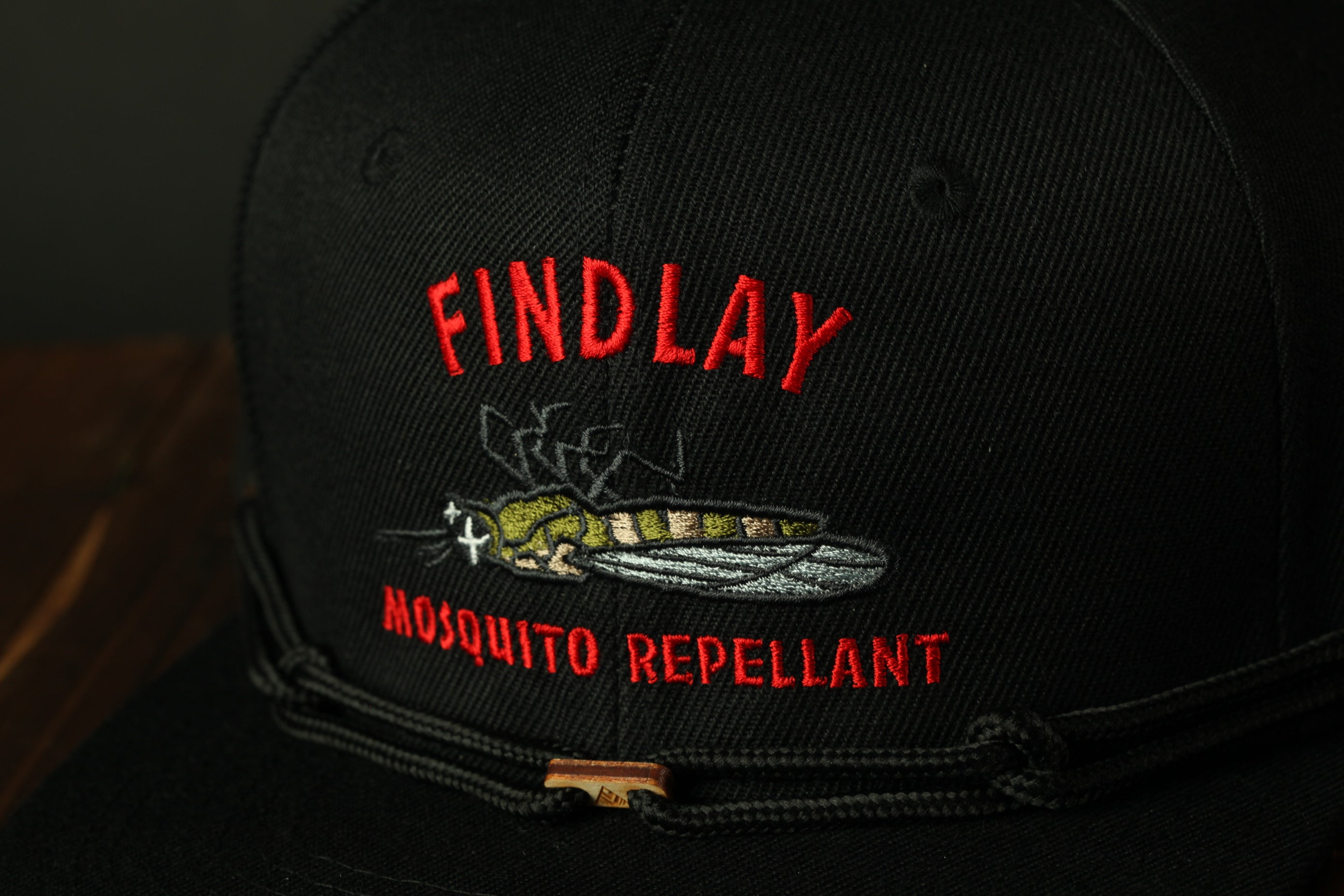 A black baseball cap with a red and white embroidered design on it, featuring a mosquito and the text "FIND LAY" and "MOSQUITO REpellent".