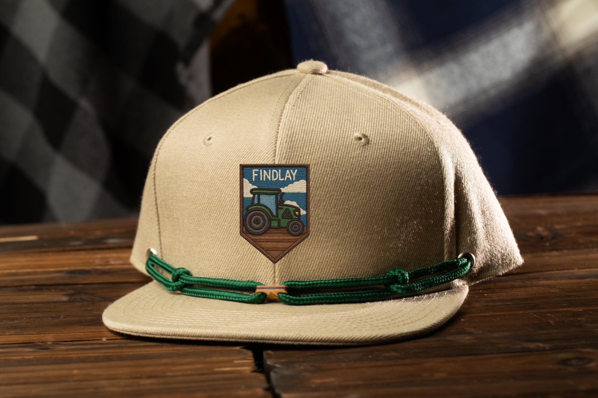A beige baseball cap with a green string and a patch featuring a tractor on it, placed on a wooden surface.