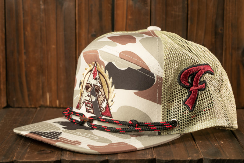 A camouflage baseball cap with a red and black embroidered logo on the side, sitting on a wooden surface.