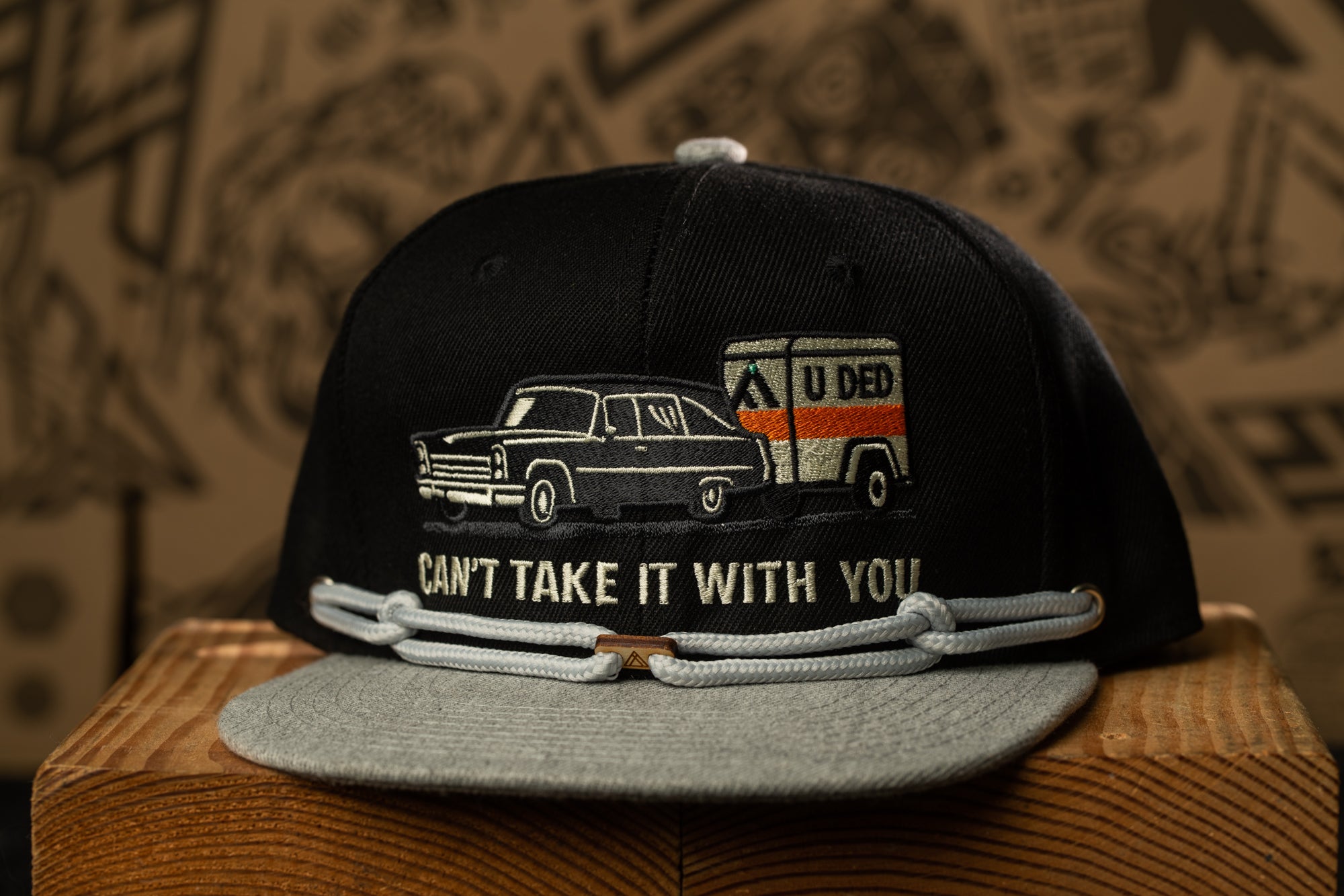 A black baseball cap with a graphic design on it, featuring a car and a trailer, along with the text "CAN'T TAKE IT WITH YOU".