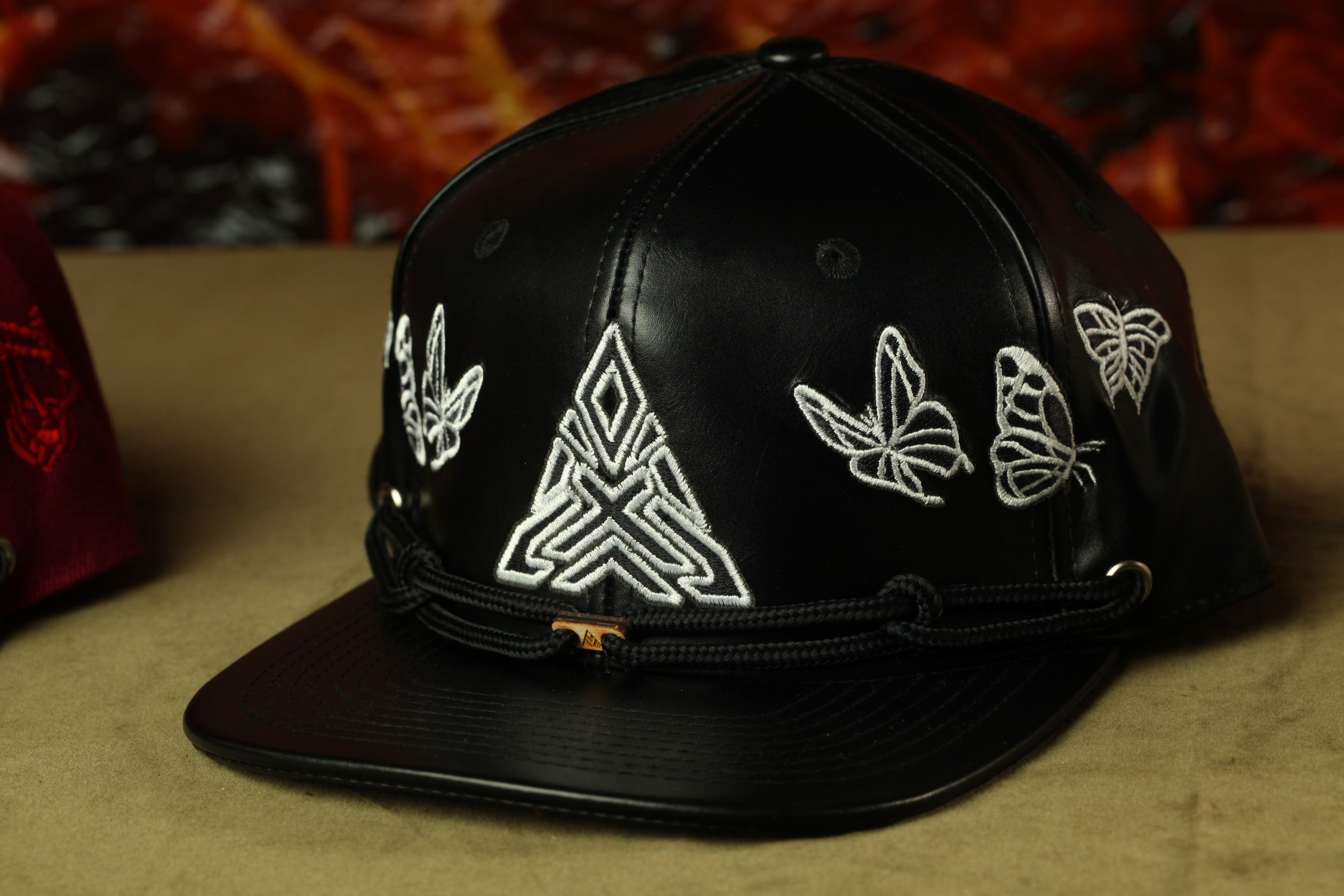 A black baseball cap with white embroidered designs, including butterflies and geometric patterns, is displayed on a surface.