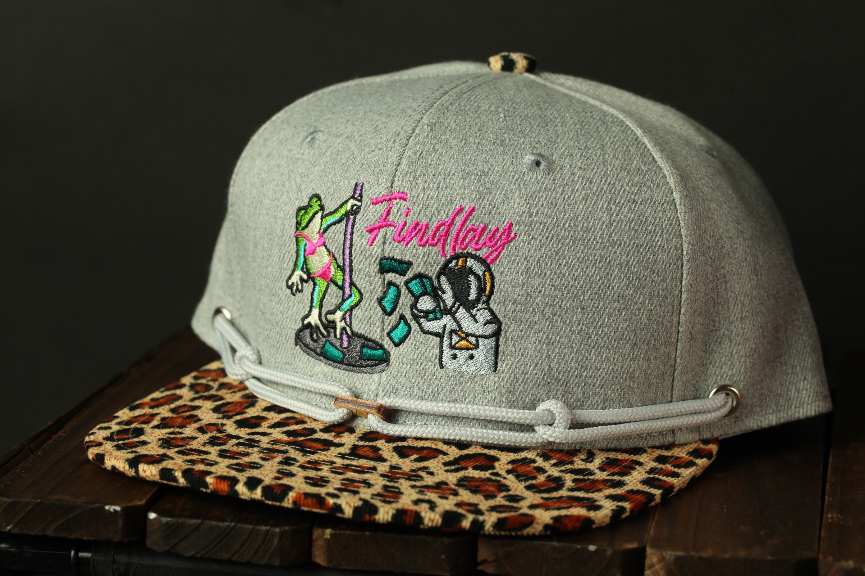 A grey baseball cap with a leopard print band and a graphic of a frog and a person on it, along with the word "Findlay" printed on it.