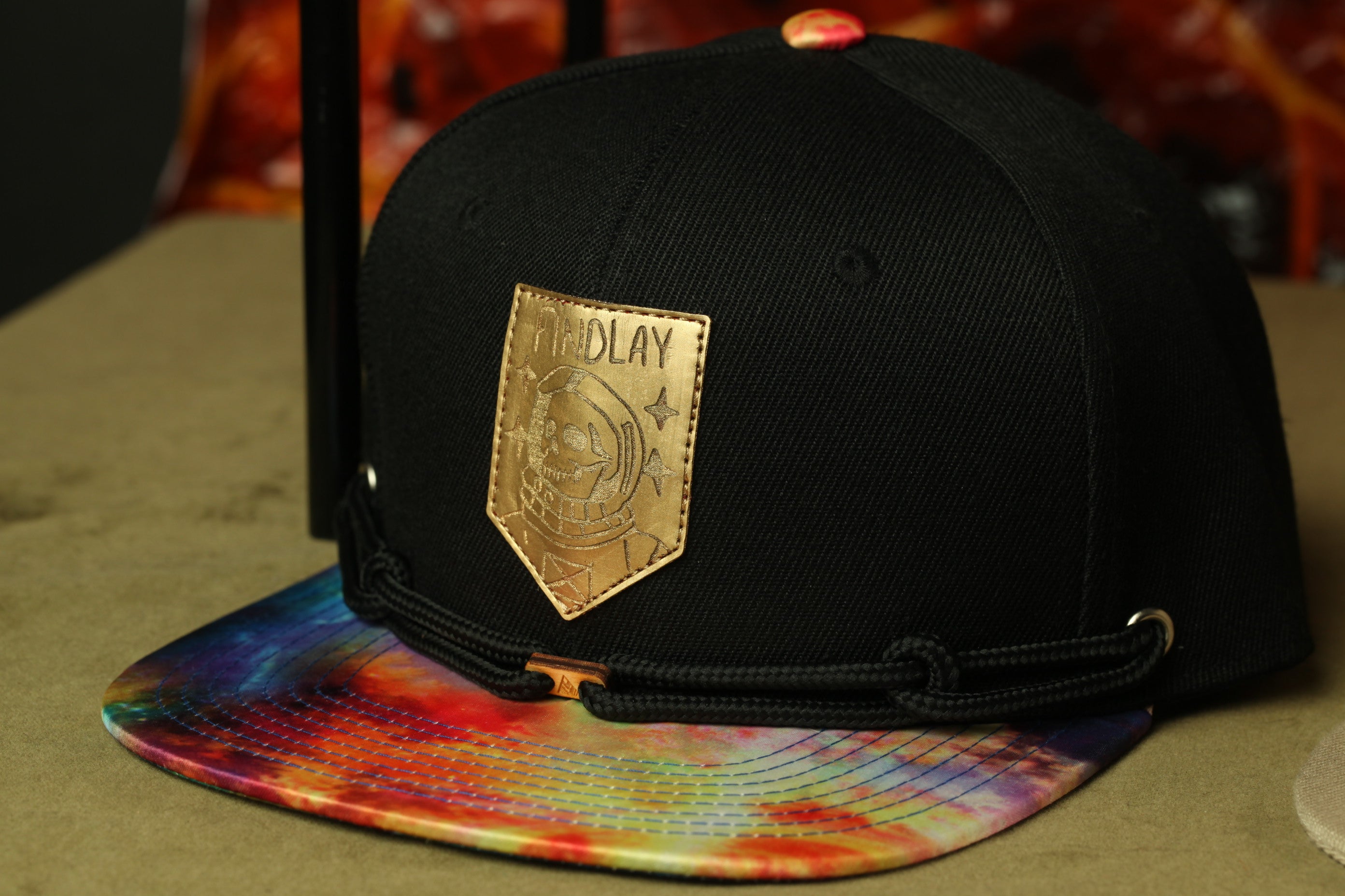 A black baseball cap with a gold patch on the front, featuring a colorful tie-dye pattern on the brim.