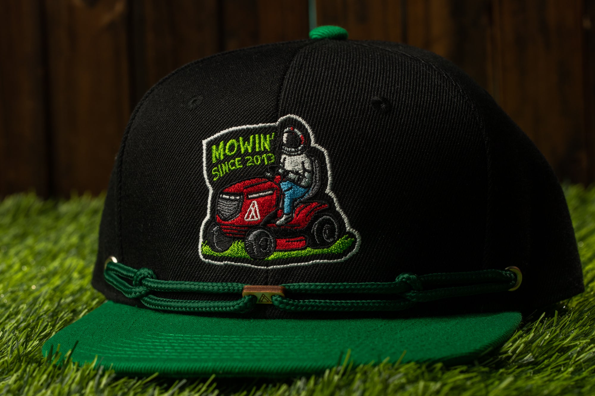 A black baseball cap with a green brim, featuring an embroidered design of a person riding a red lawn mower.