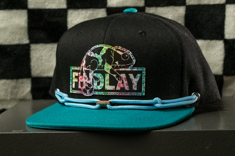 A black baseball cap with a colorful graphic and the word "FIND LAY" printed on it, sitting on a black surface against a checkered background.