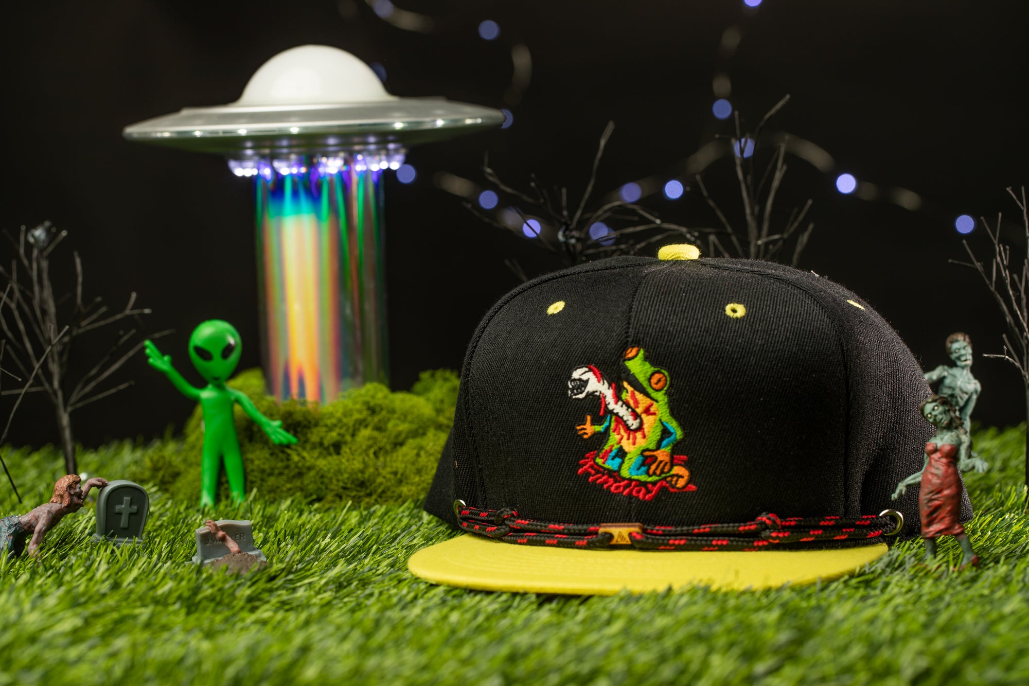 a black baseball cap with a colorful design, surrounded by various toys and figurines, including a green alien figure, a green monster, and a green zombie. The scene is set against a backdrop of a green grassy area with a spaceship and a glowing alien structure.