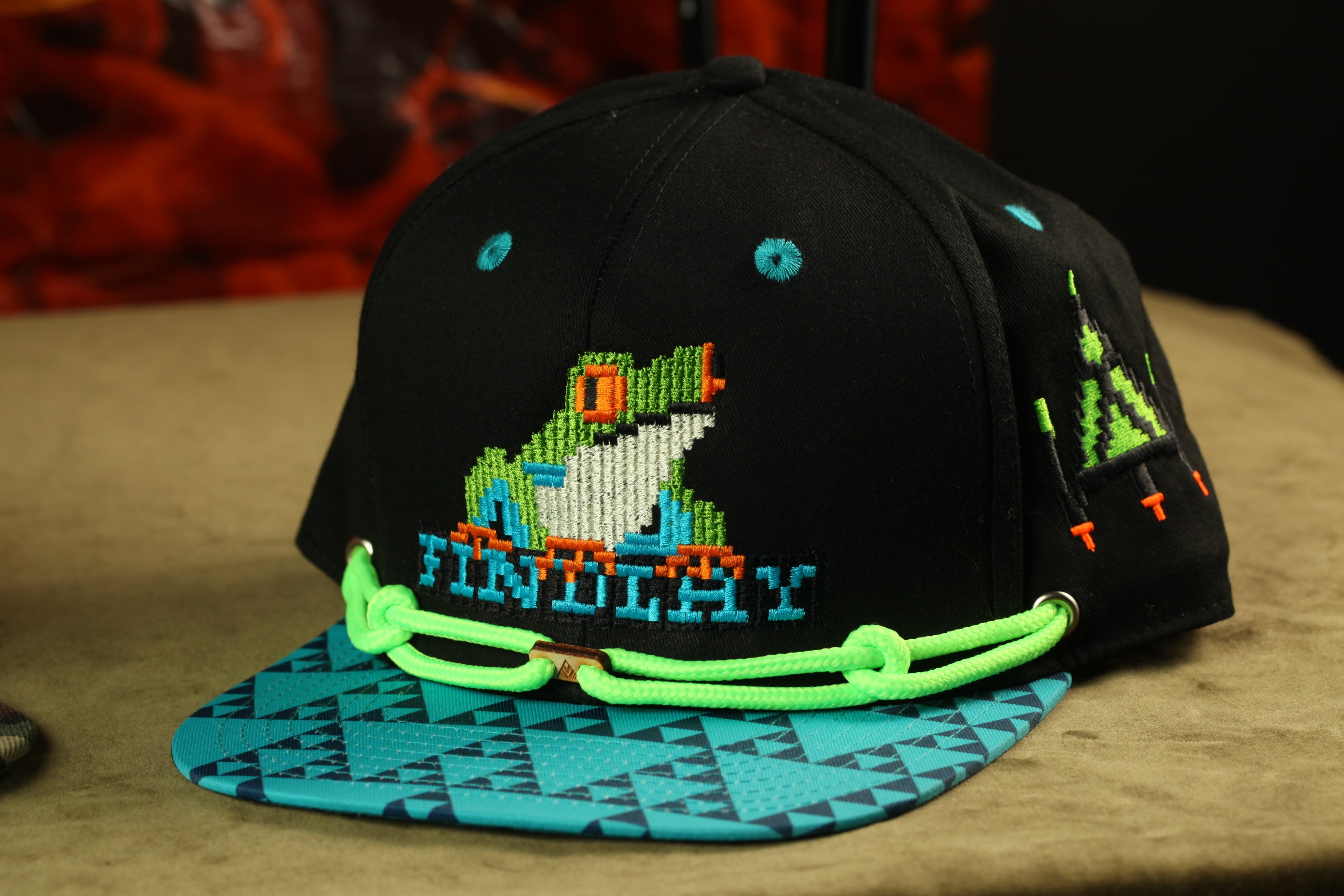 A black baseball cap with a colorful, pixelated design of a frog on the front, and a geometric pattern on the brim.