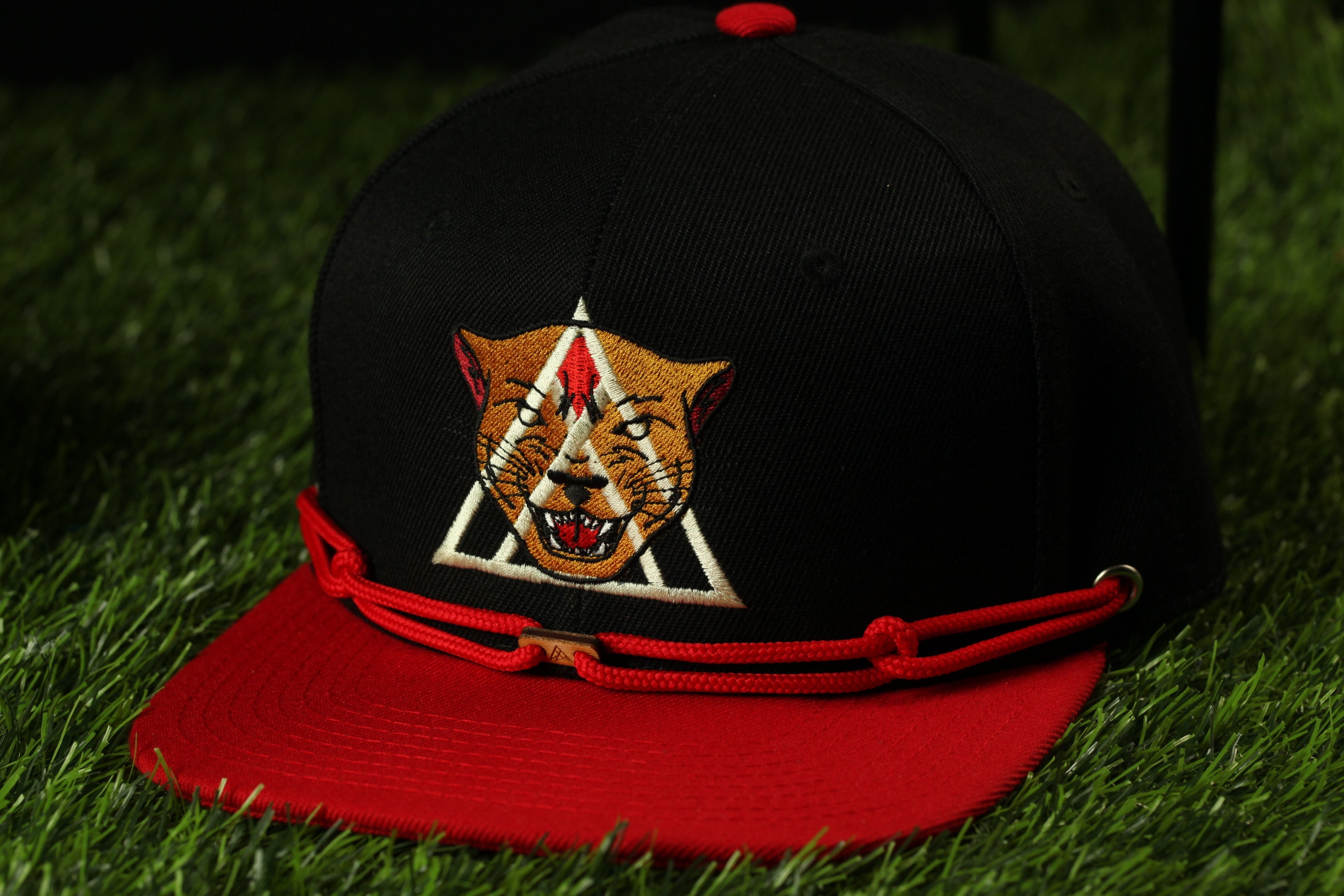 A black baseball cap with a red brim and a graphic of a cat's face on the front, placed on a grassy surface.