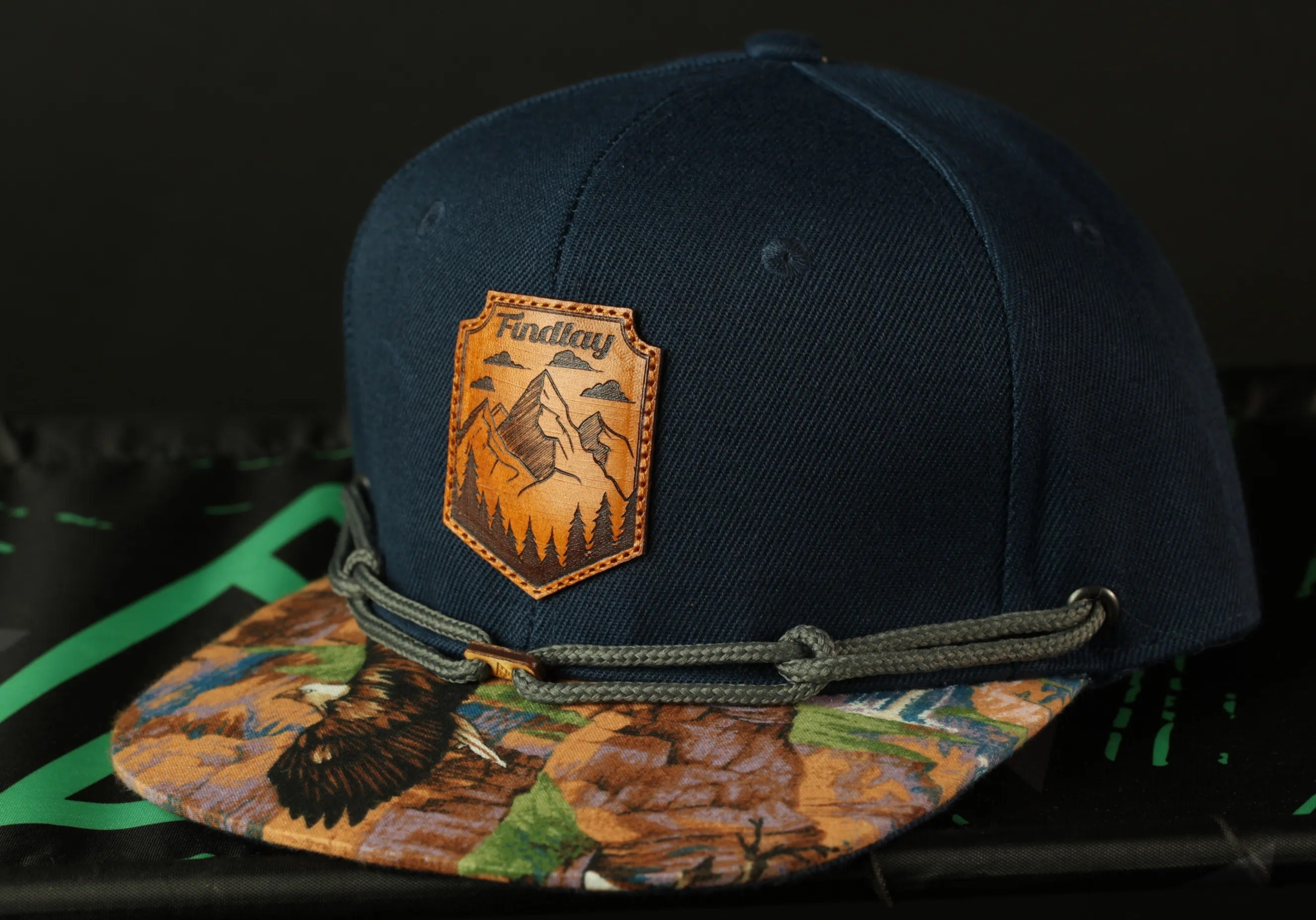 A navy blue baseball cap with a mountain-themed patch and a colorful printed pattern on the brim.